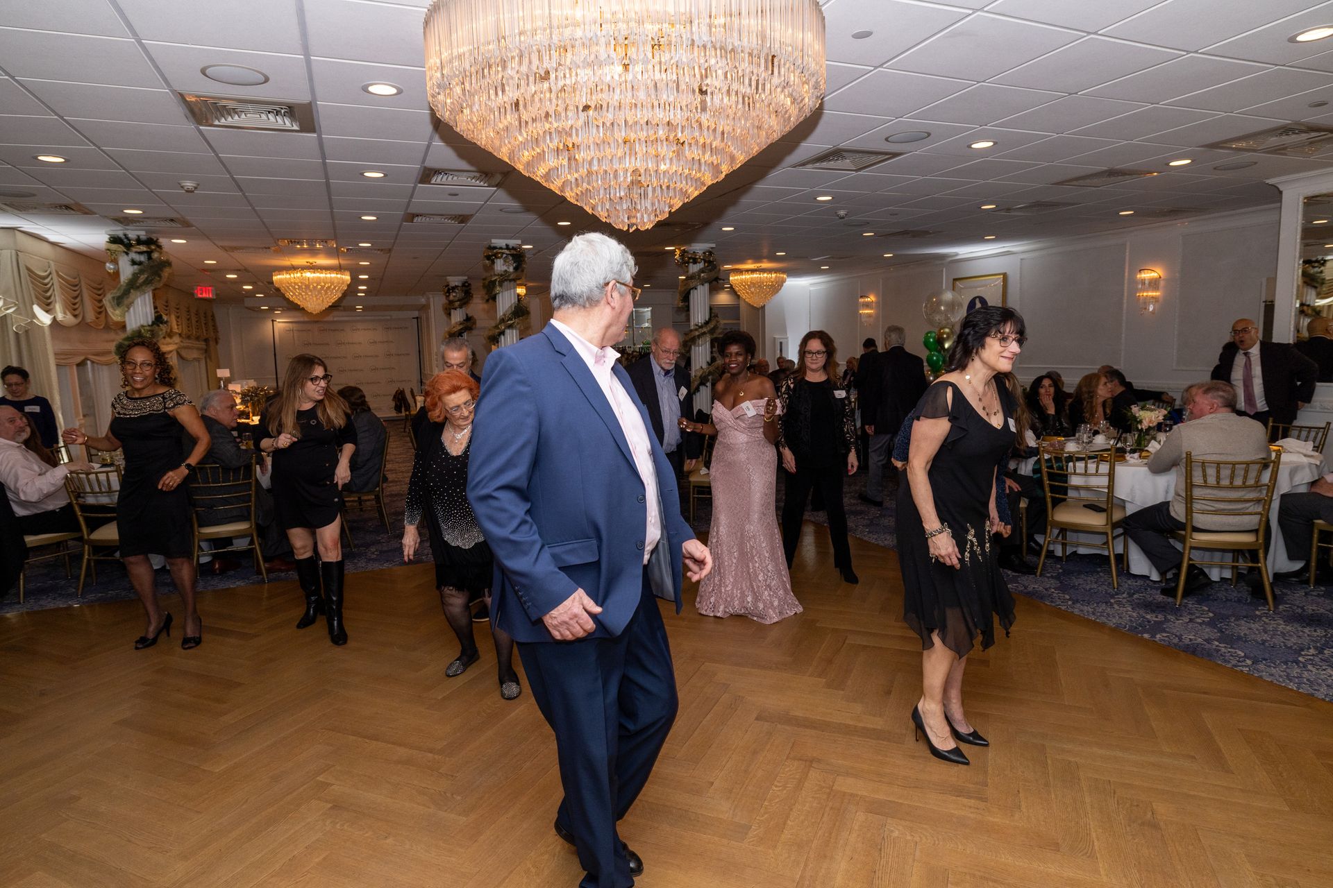 People dancing in a ballroom; chandelier, tables, and decorations in background.