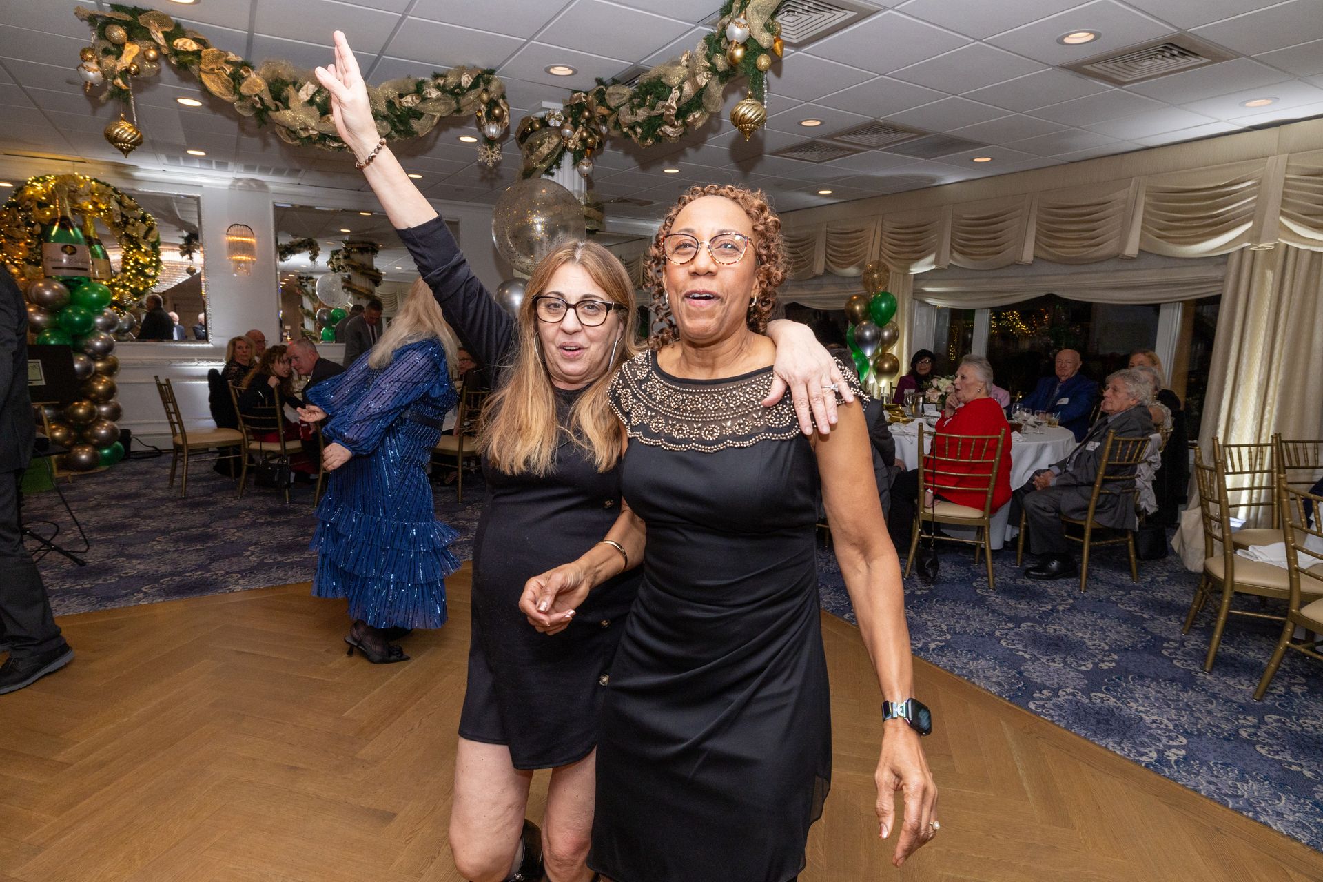 Two women dancing joyfully at a party, arm in arm. Decorated room.