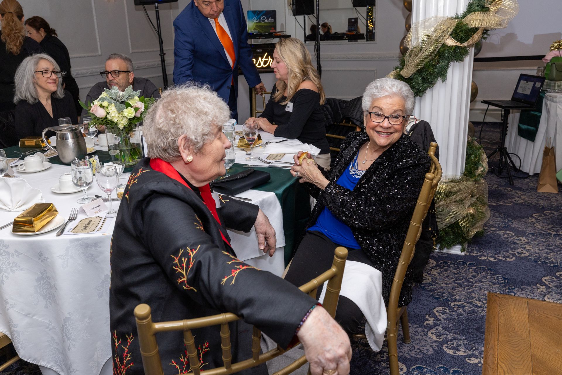Two women at a table, smiling, in a decorated dining room. Other people are seated at tables.