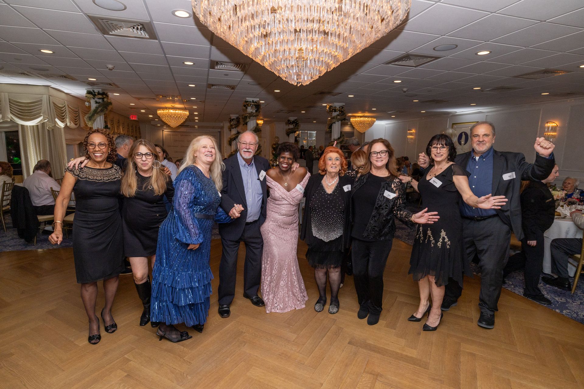 Group of people dancing in a ballroom, under a chandelier. They are smiling and having fun.