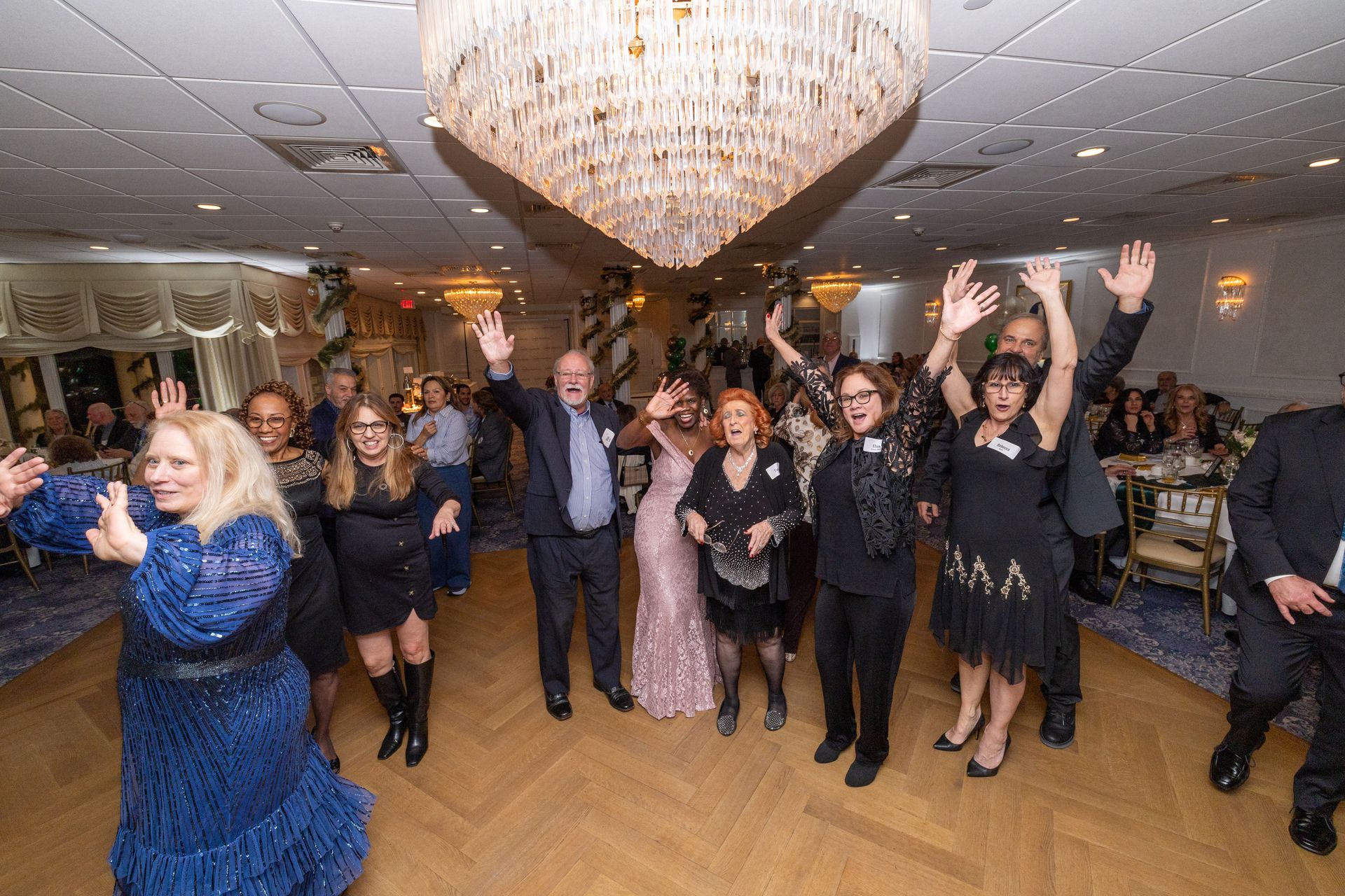 People dancing with arms raised in a brightly lit ballroom, a chandelier overhead.