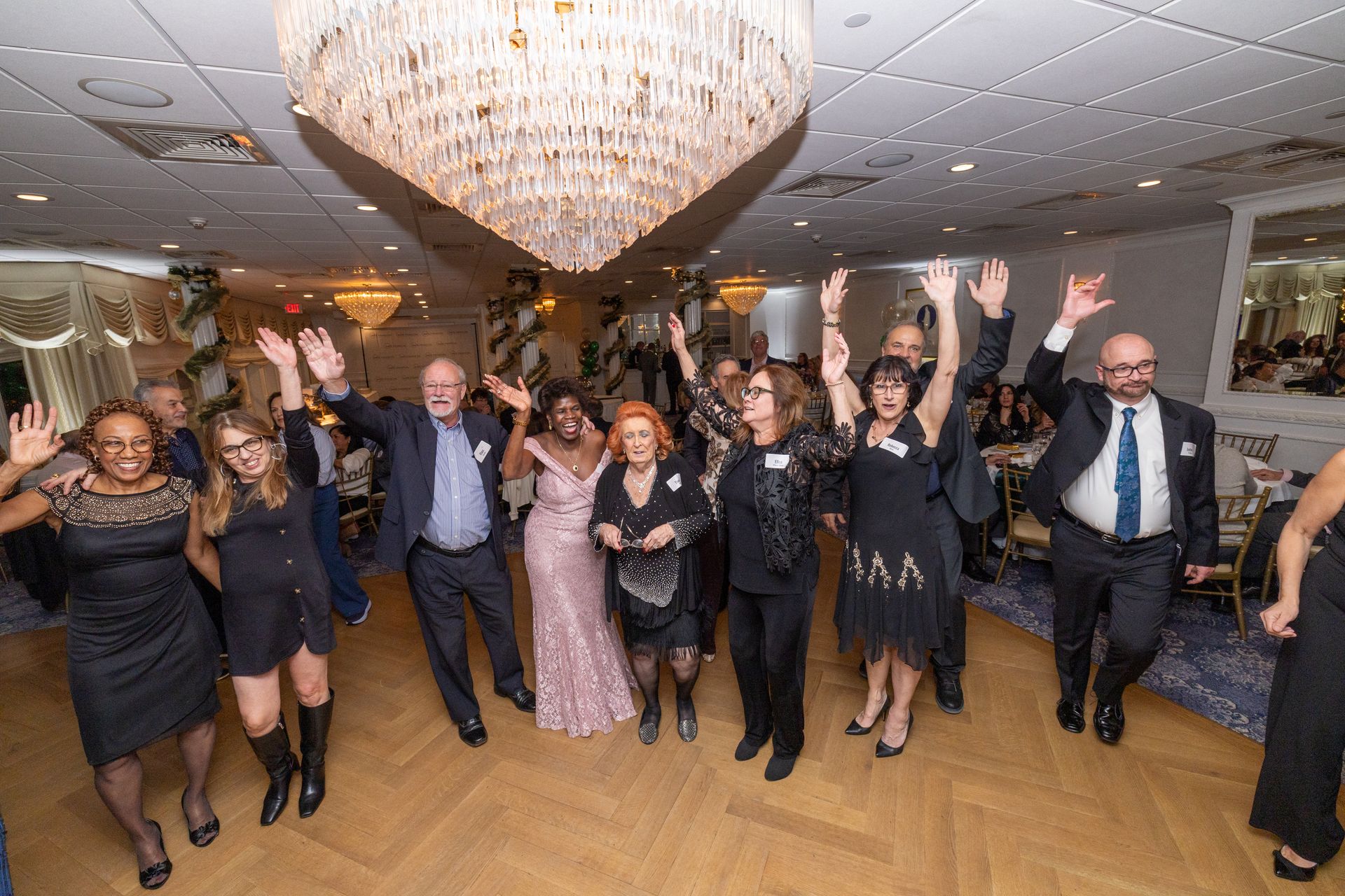 Group of people celebrating in a banquet hall, arms raised. Elegant chandelier overhead.