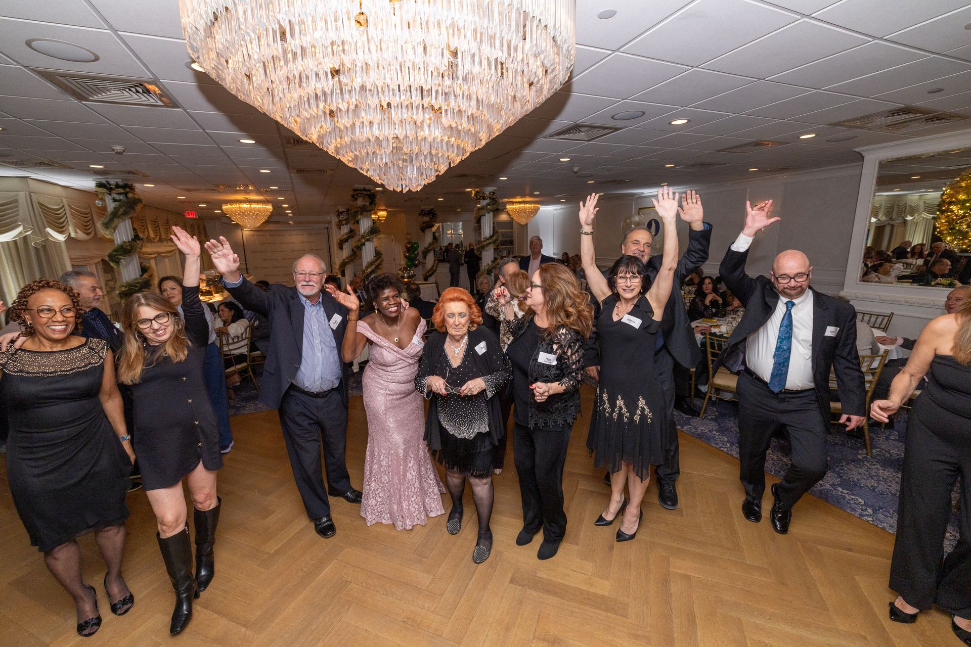 People celebrating at an event in a ballroom with a large chandelier. Several have arms raised, smiling.