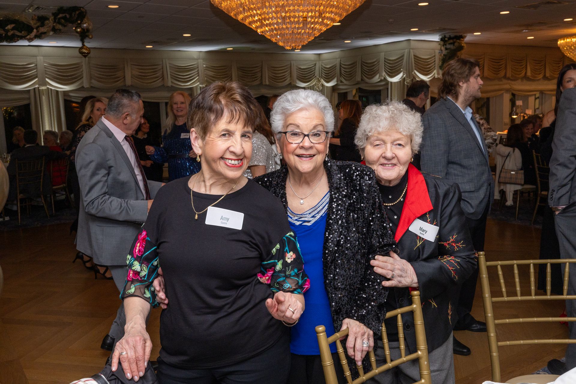 Three older women smiling and posing together at an event. They are standing arm-in-arm in a banquet hall.