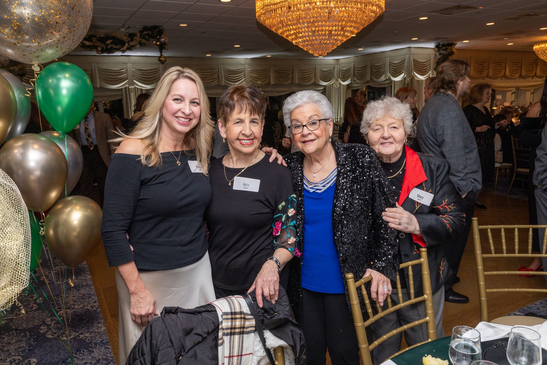 Four women smiling at an event; gold and green balloons, chandelier, formal attire.