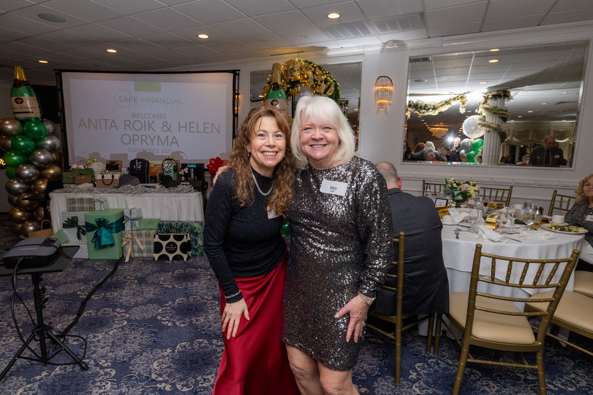 Two smiling women at an event, one in a sequined dress, standing by a gift table and a projected screen.