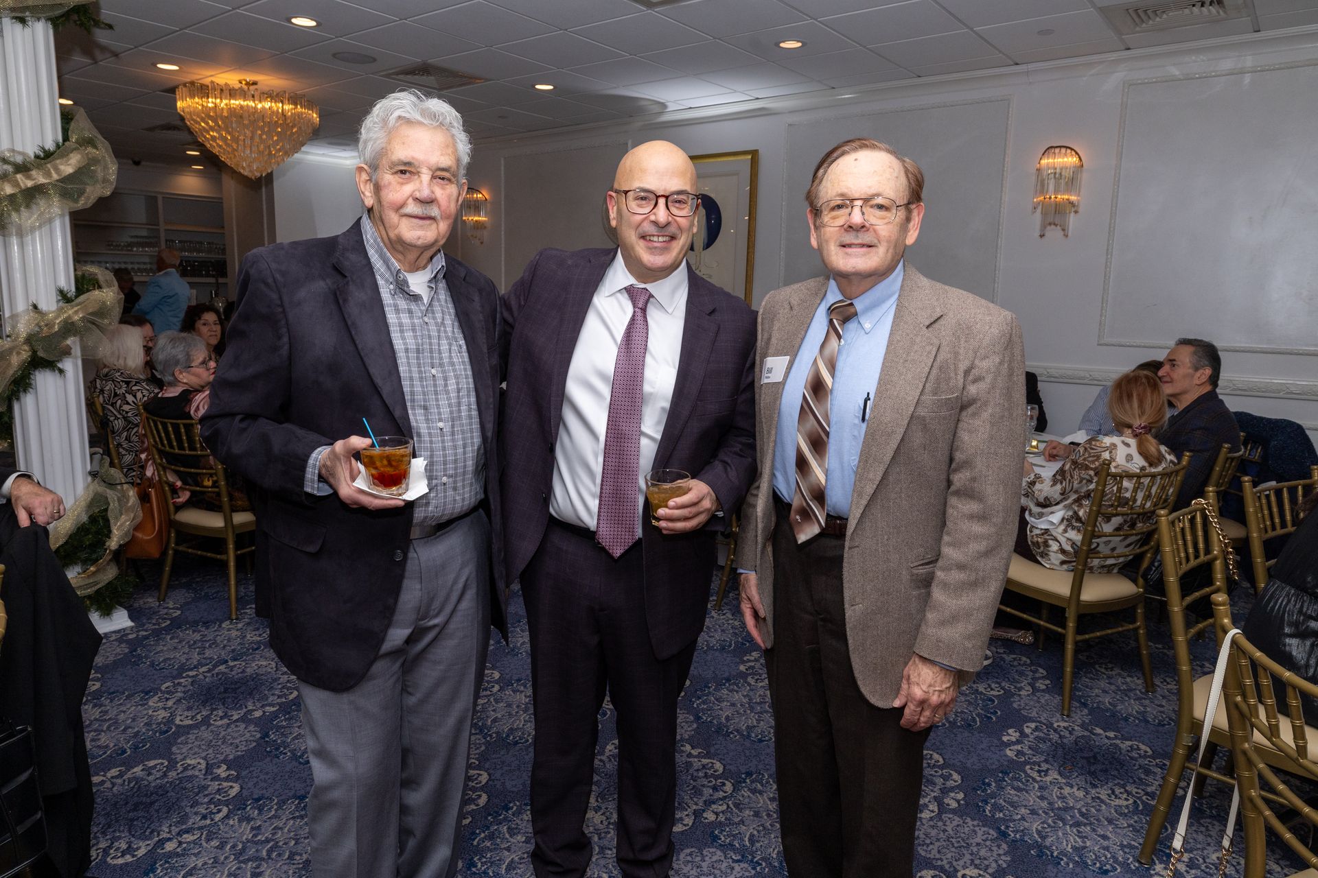 Three men in suits smiling, holding drinks, indoors with party decorations.