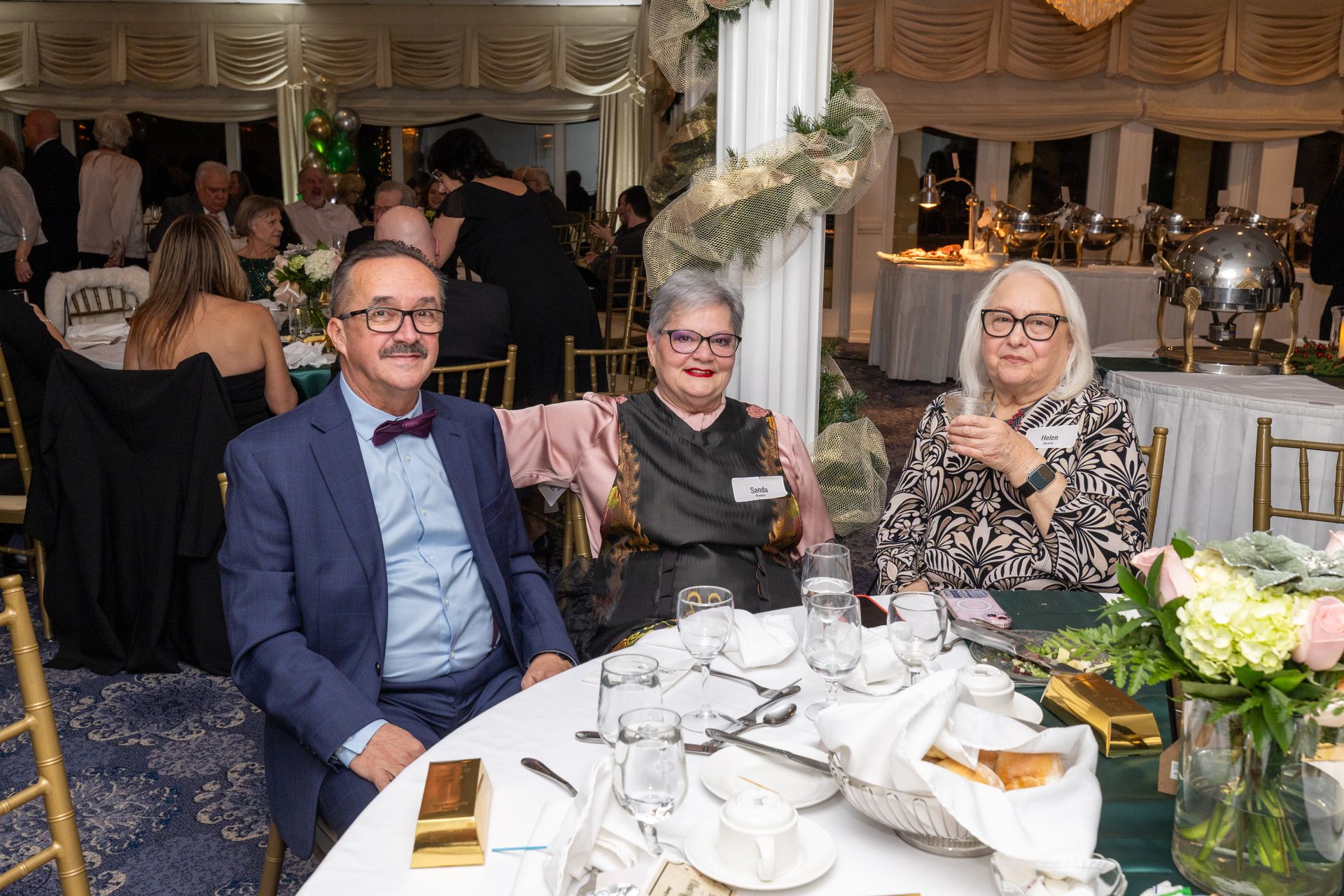 Three people at a formal event, seated at a table. Man in a suit with glasses, two women, festive decor.