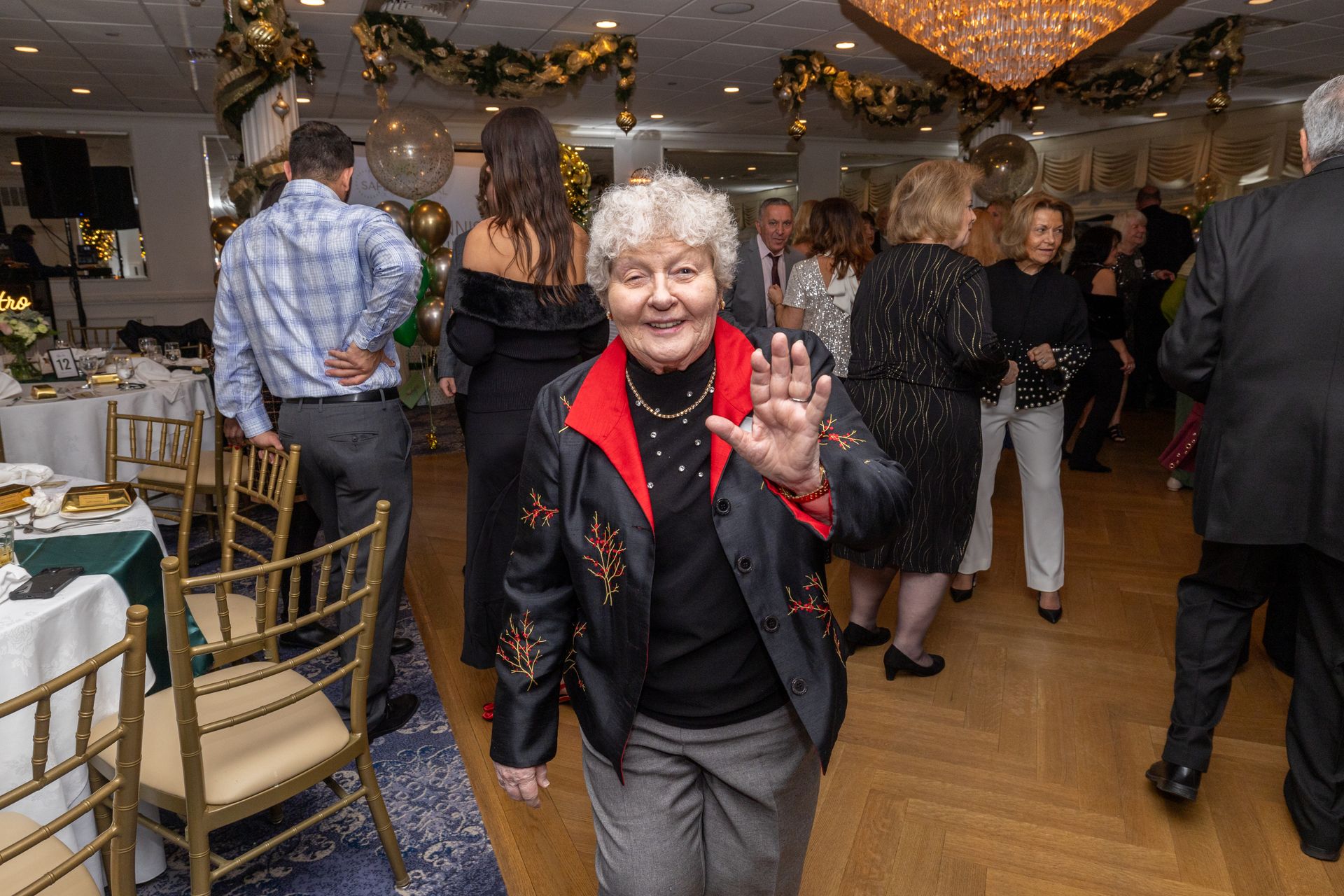 Woman waving, smiling, at a party, other people in the background, festive decorations.