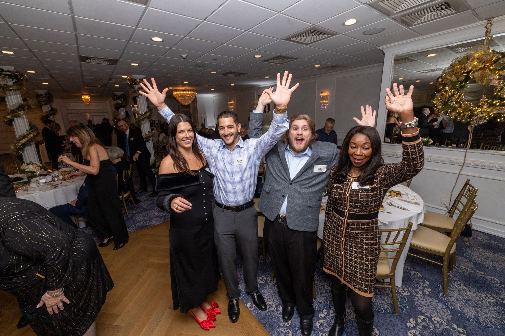 Four people cheering, arms raised, at a decorated event with tables and a chandelier.