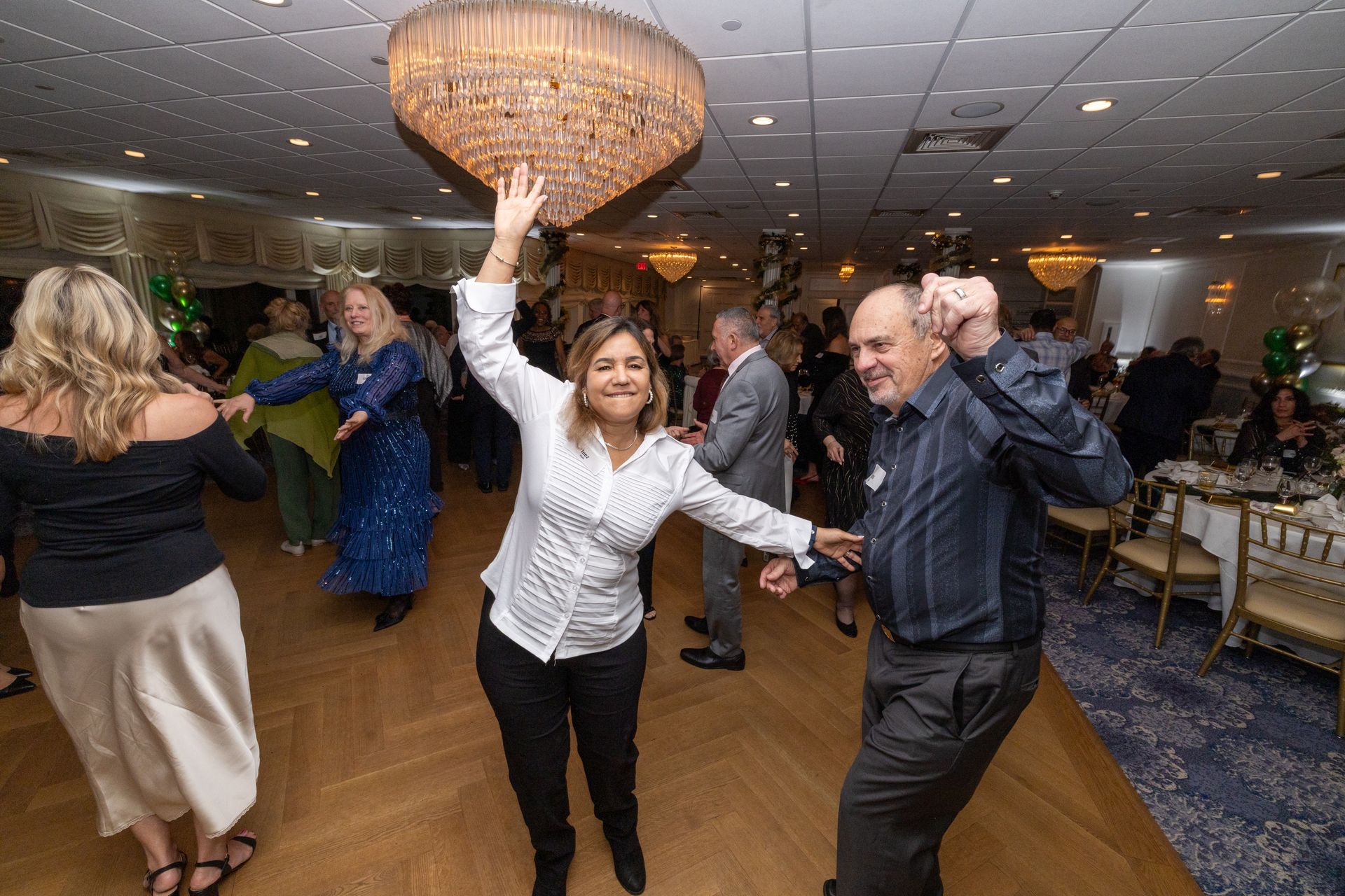 People dancing in a brightly lit ballroom. A woman raises her arms, a man smiles.