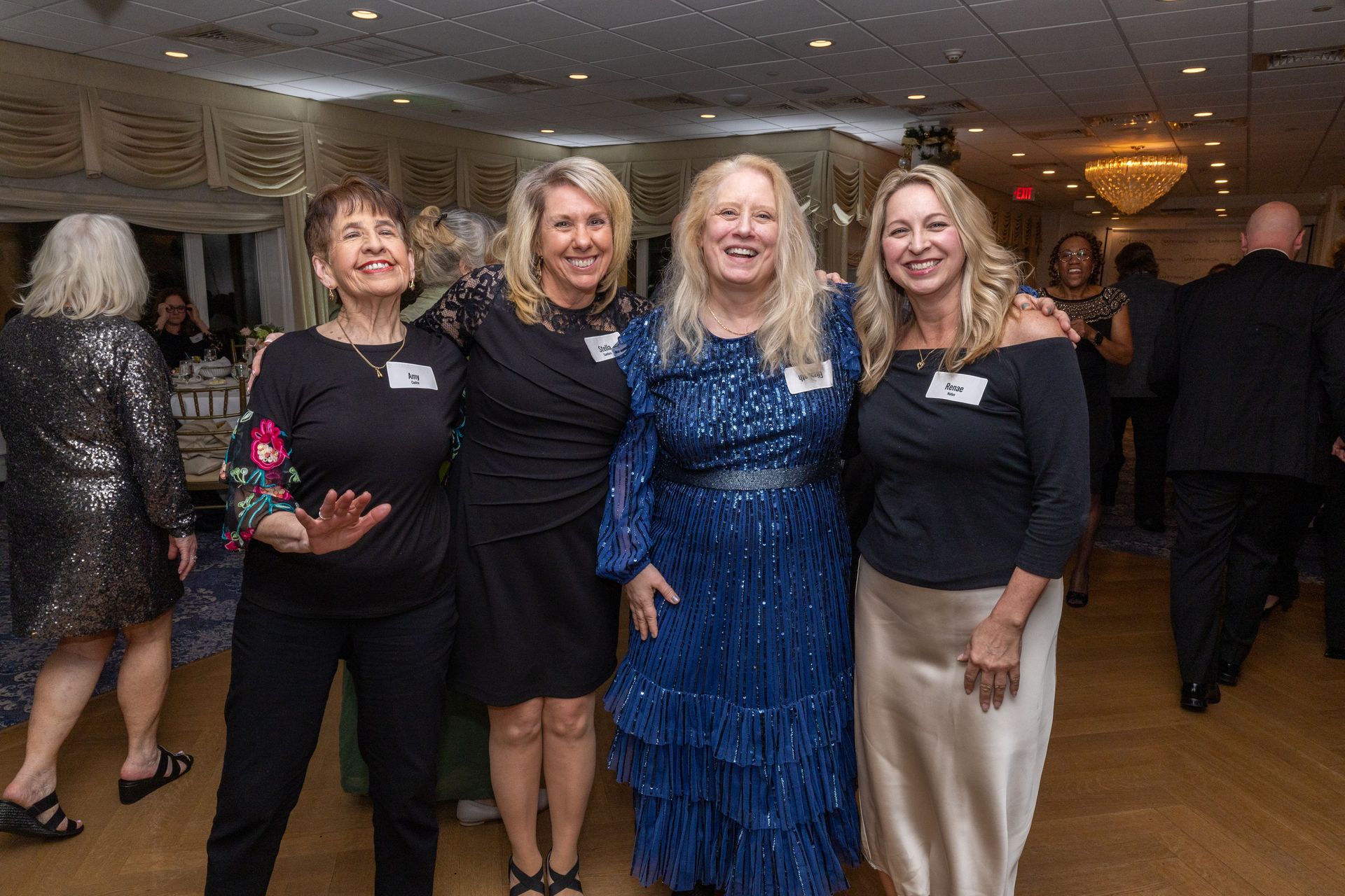 Four women pose for a photo at a formal event, smiling, in a ballroom with a disco ball.