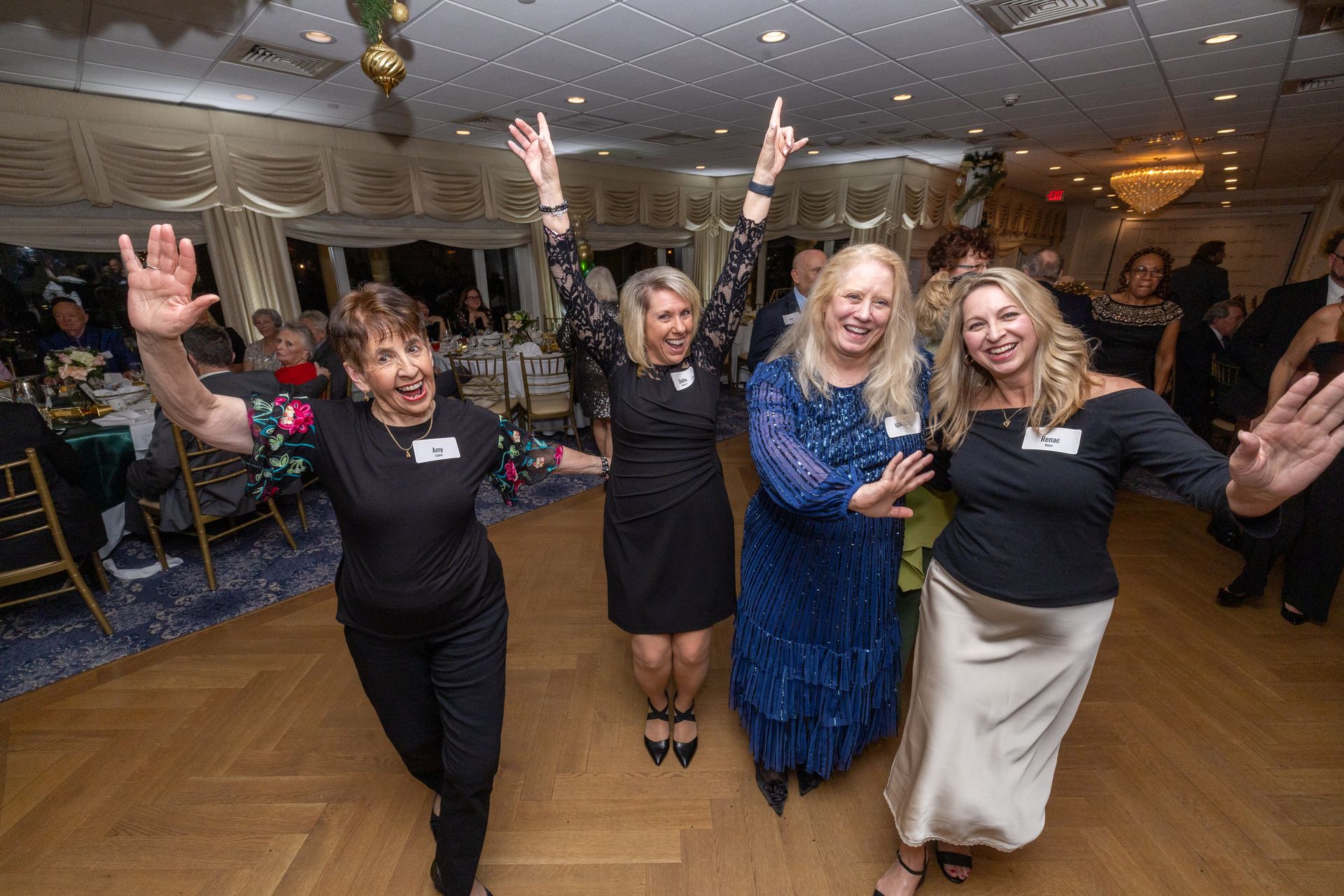 Four people dancing joyfully at an event; arms raised, smiles, in a ballroom with decor.