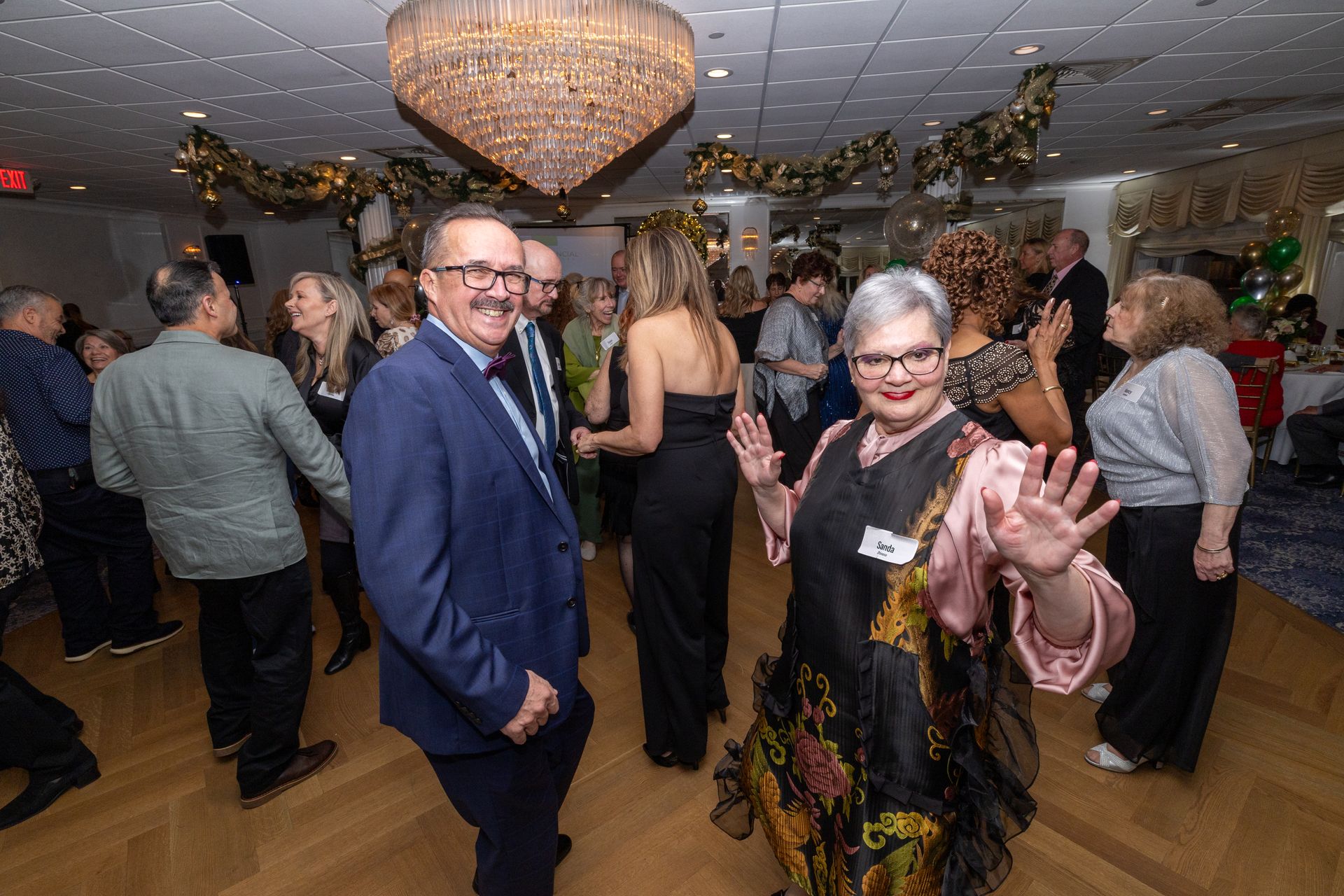 People dancing at a party under a chandelier; many wearing formal attire.