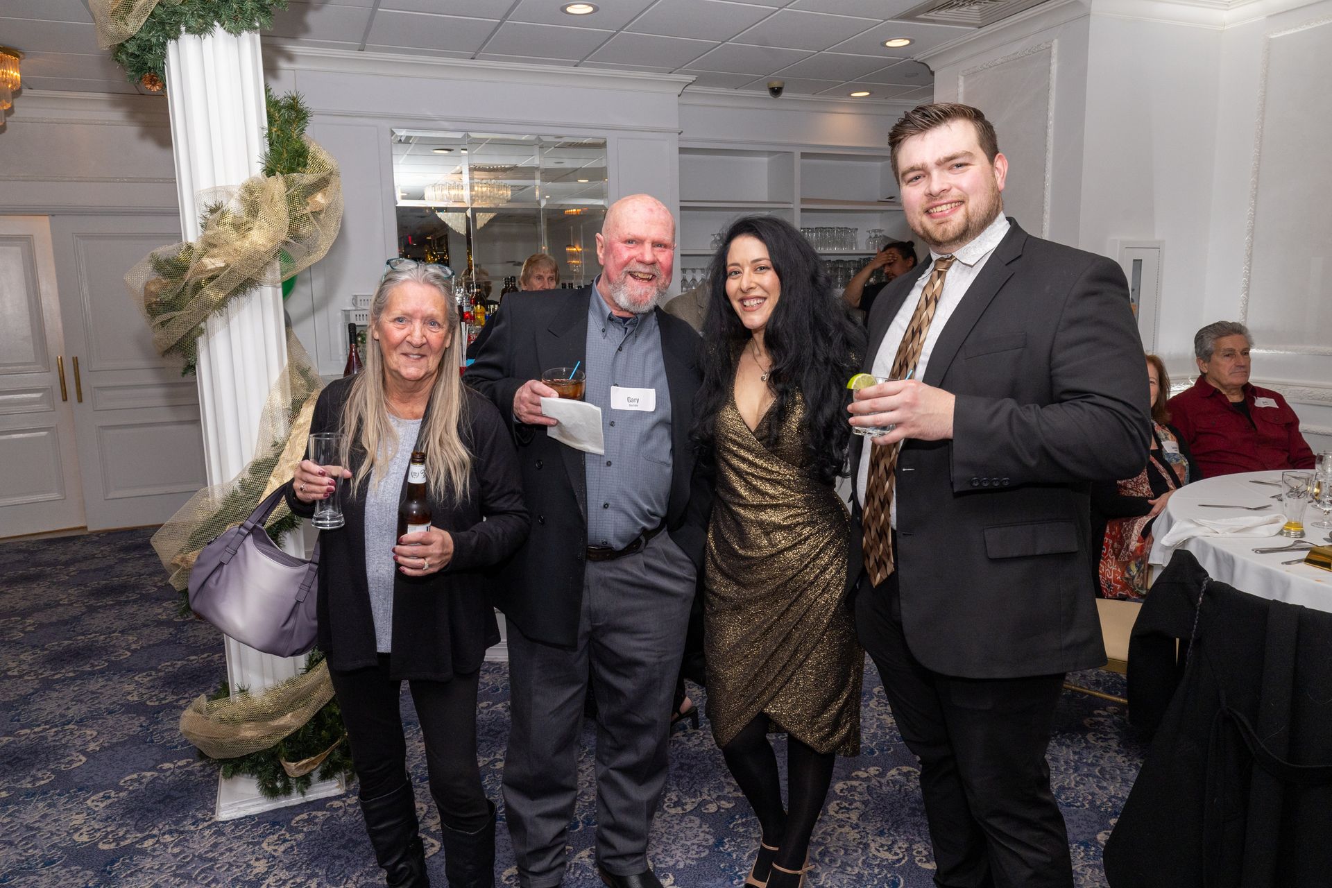 Four people pose at an event: a woman in black, a man holding a drink, a woman in a gold dress, and a man in a suit.