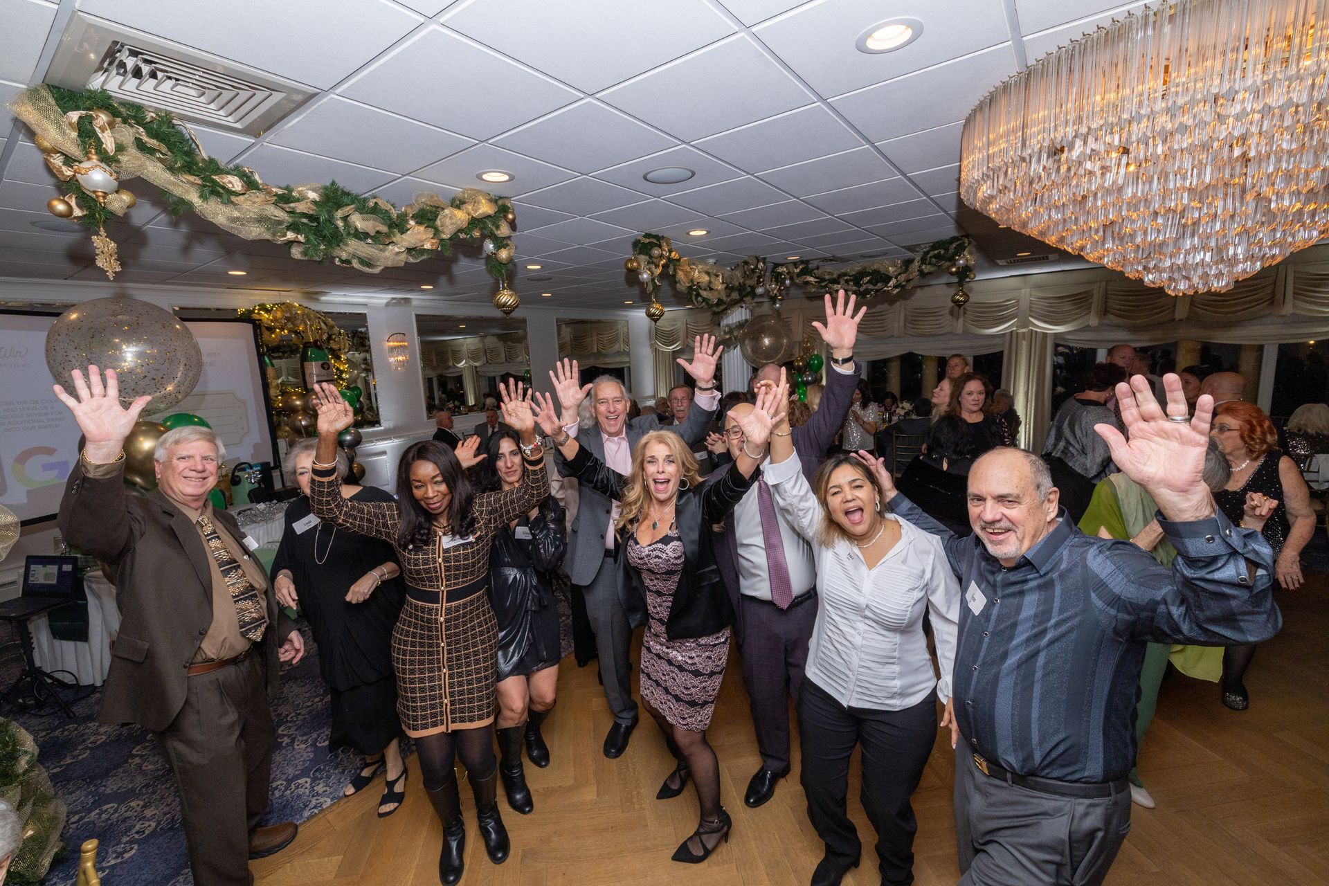 Group of people at a party, raising hands and smiling under decorations, festive lights.