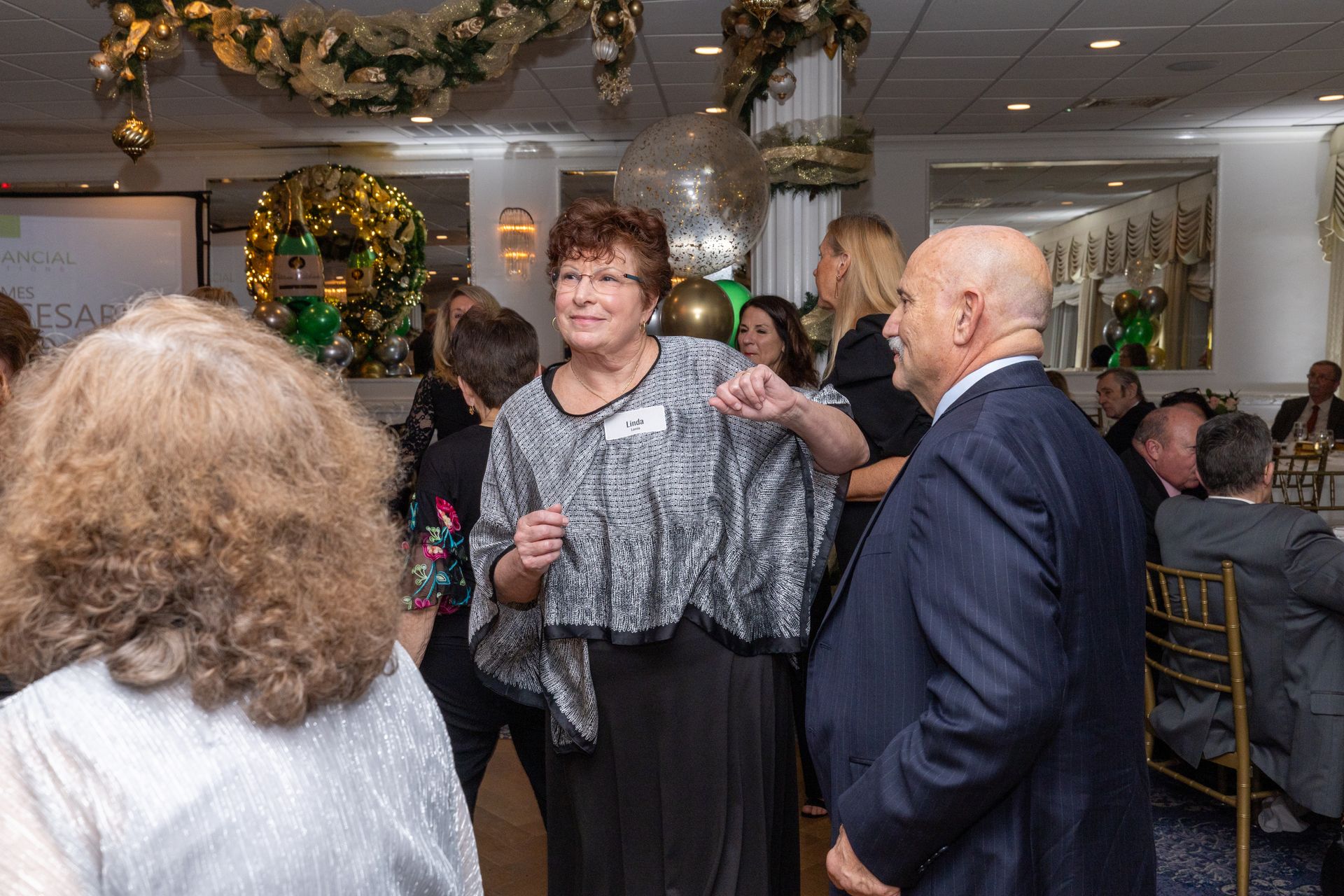 Woman dancing at a party, gesturing near a man in a suit. Gold and green decorations in a well-lit room.