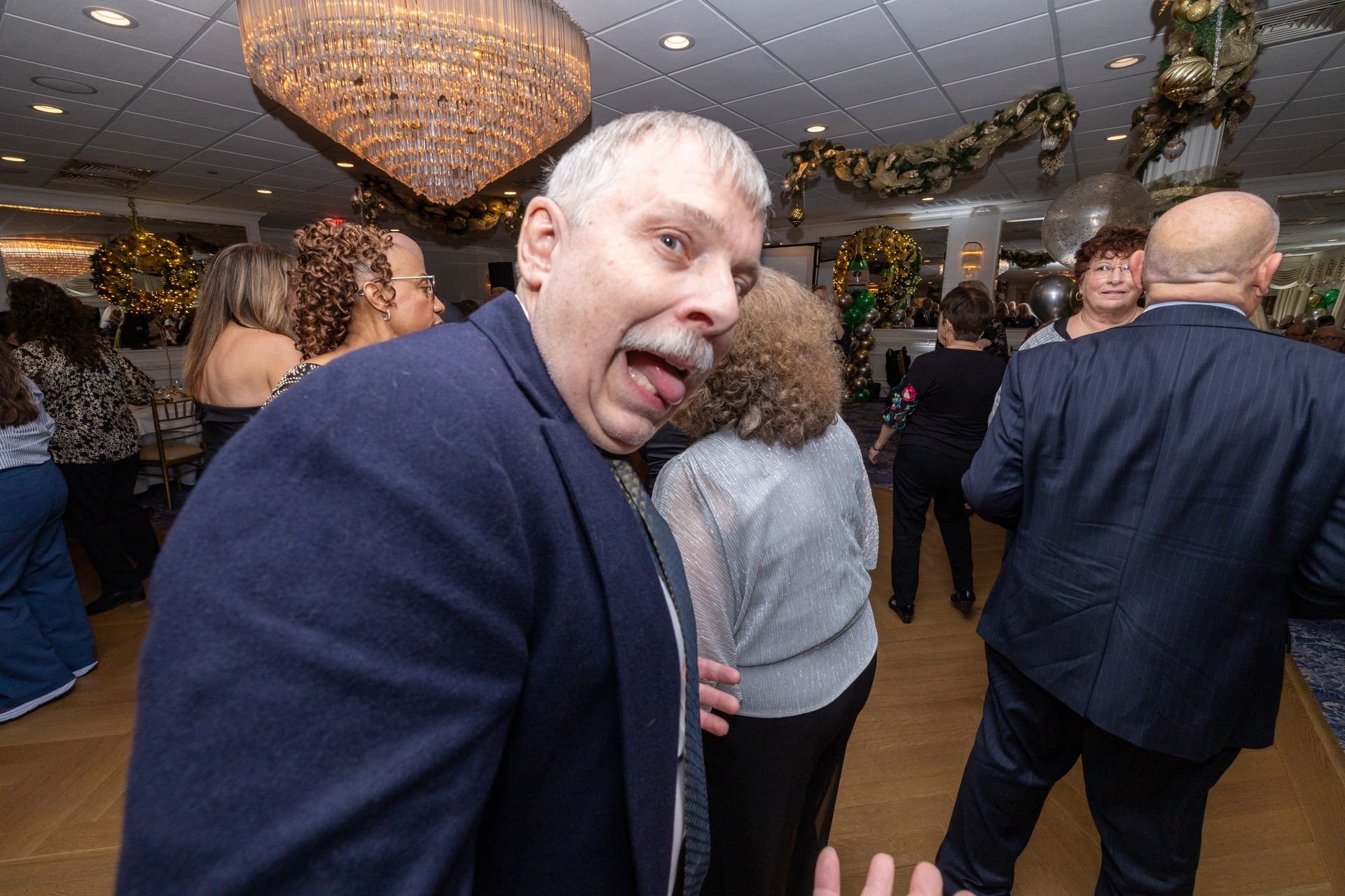 Man sticking out tongue at a party, wearing a blue jacket, dancing. Others in the background.
