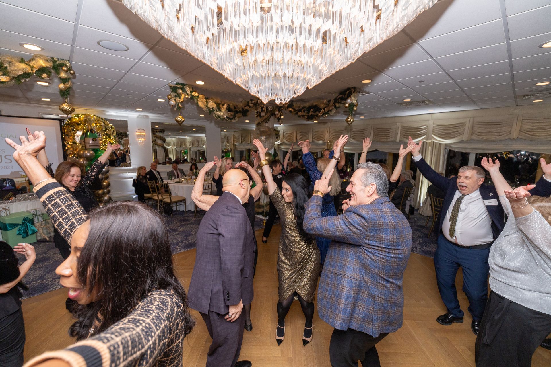 People dancing and celebrating in a brightly lit room with a chandelier. Hands raised.