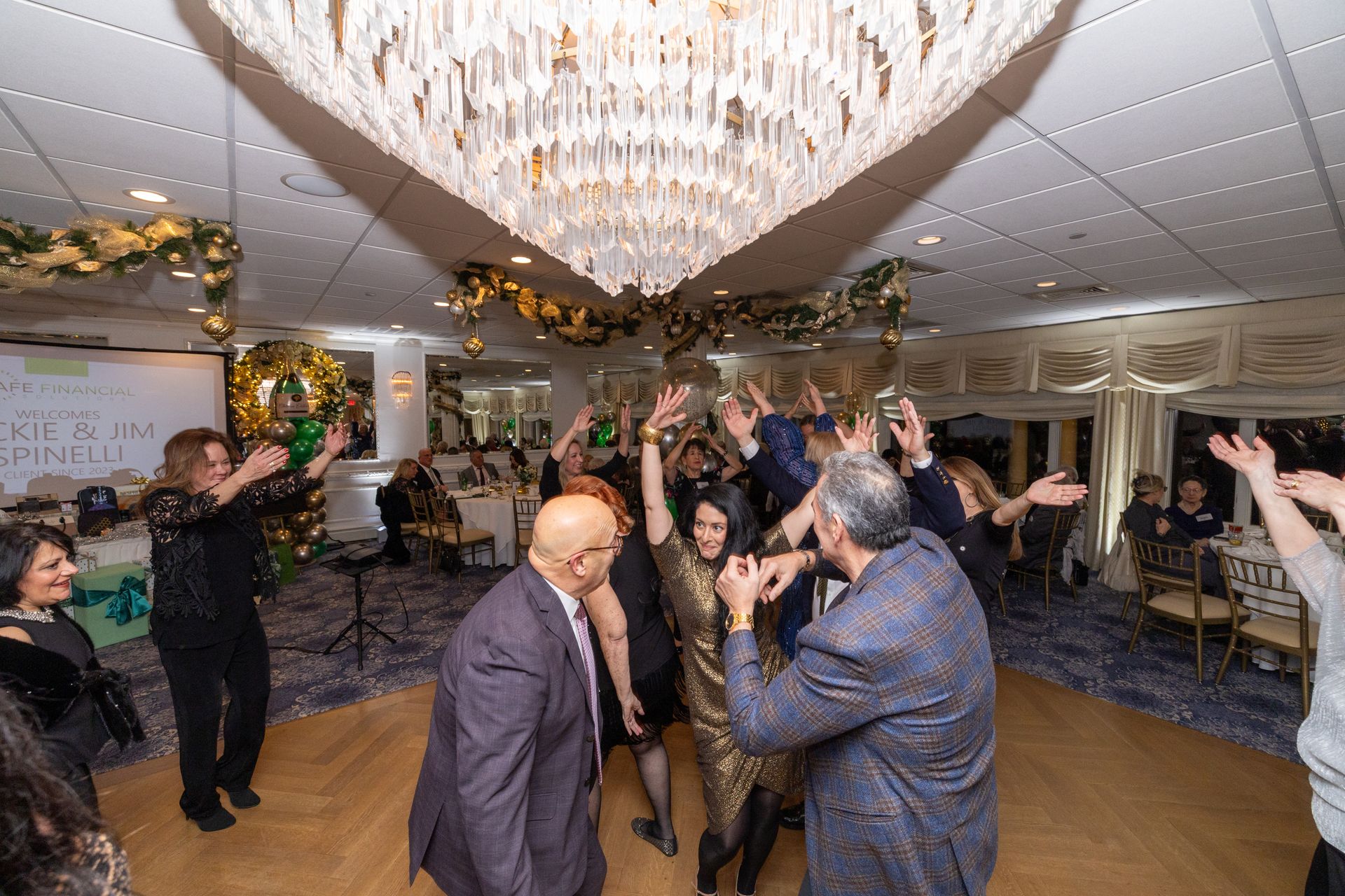 People dancing under a chandelier in a decorated ballroom. Hands raised in celebration.