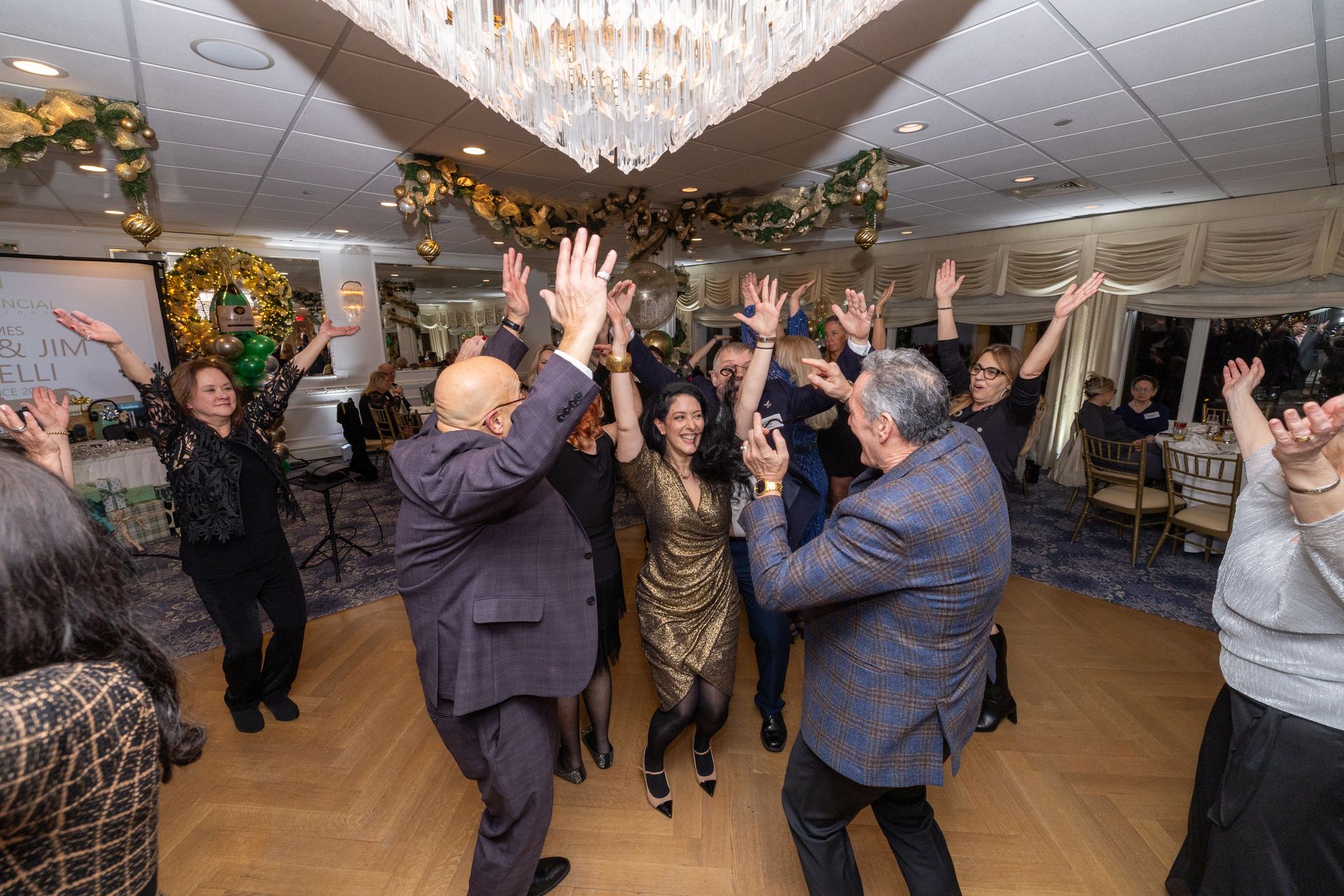 People dancing in a decorated ballroom under a chandelier, raising their arms.