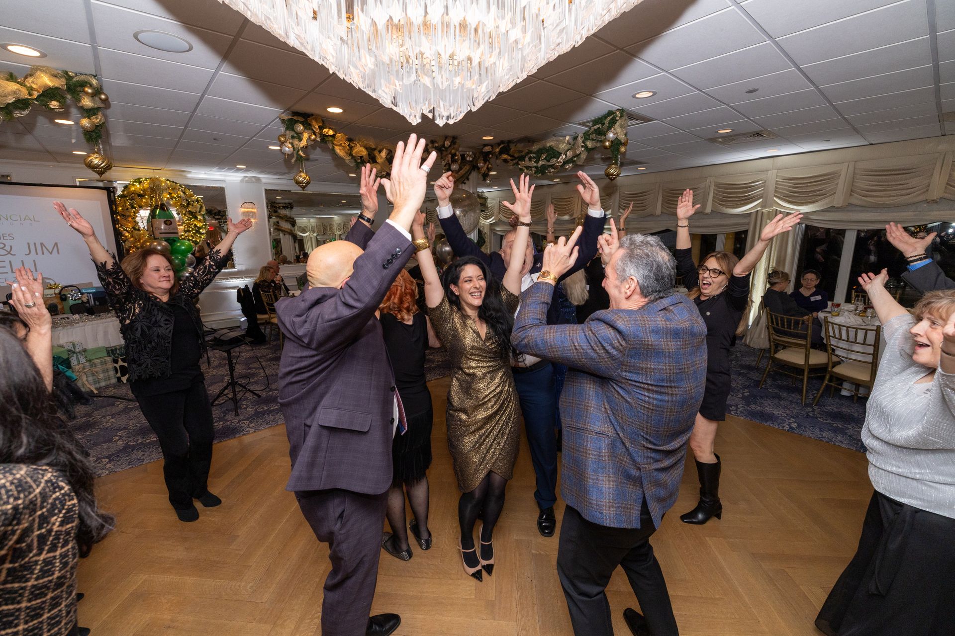 People dancing with arms raised under a chandelier in a decorated ballroom.