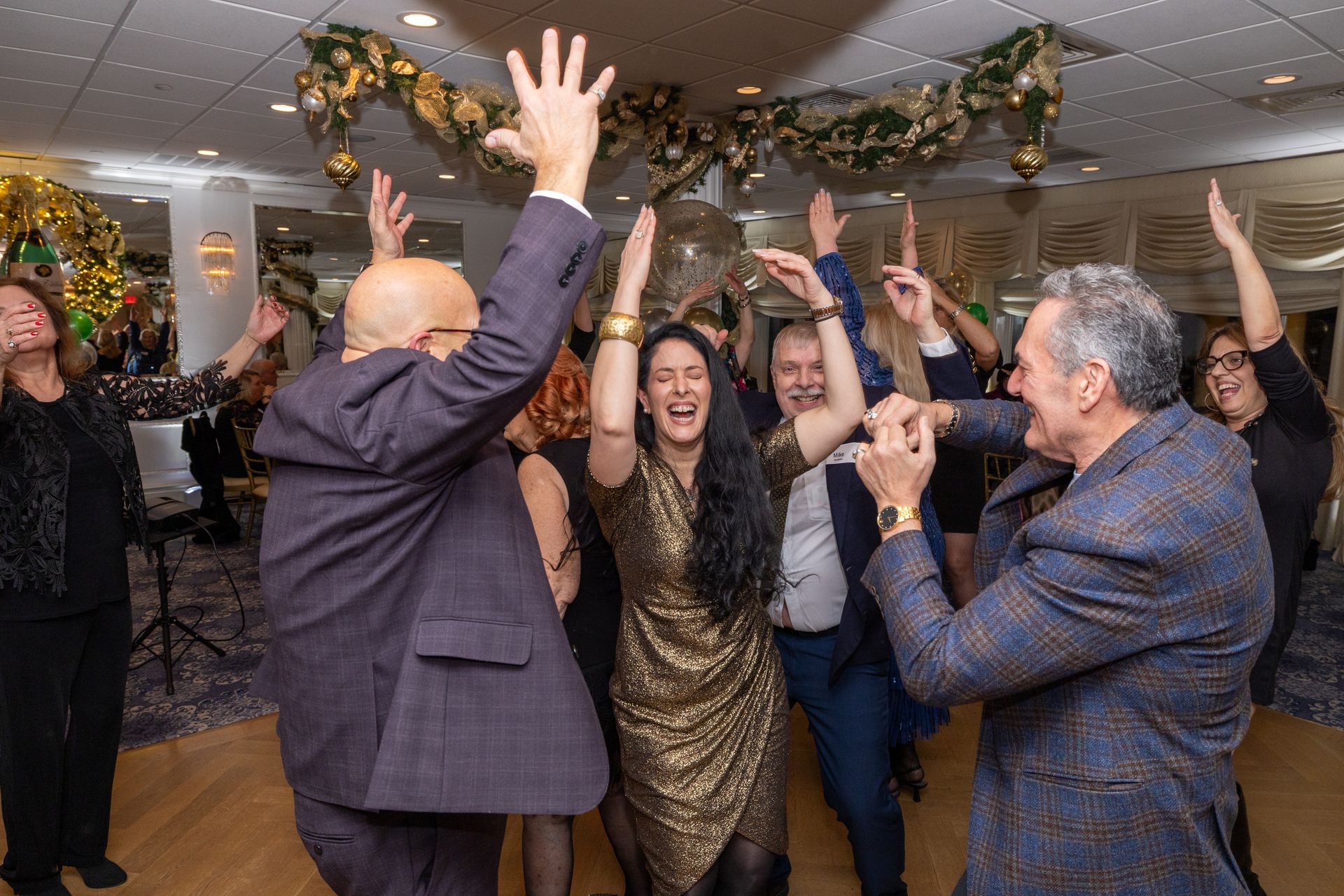 People dancing and celebrating at a festive gathering. Arms raised, joyful expressions. Floral decor overhead.