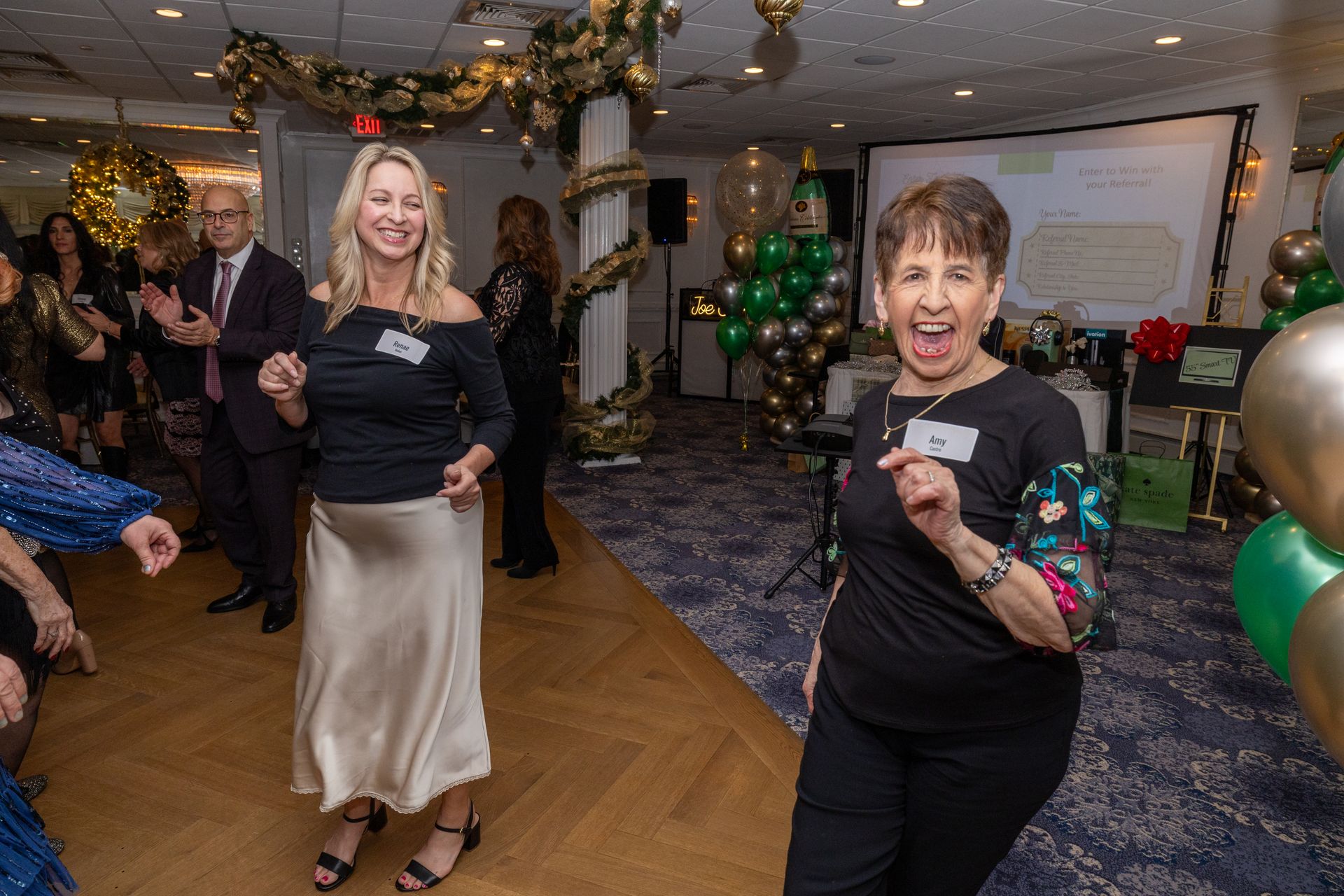 Two women dancing at an event, one in black with a gold skirt, the other in black pants. Balloons in background.