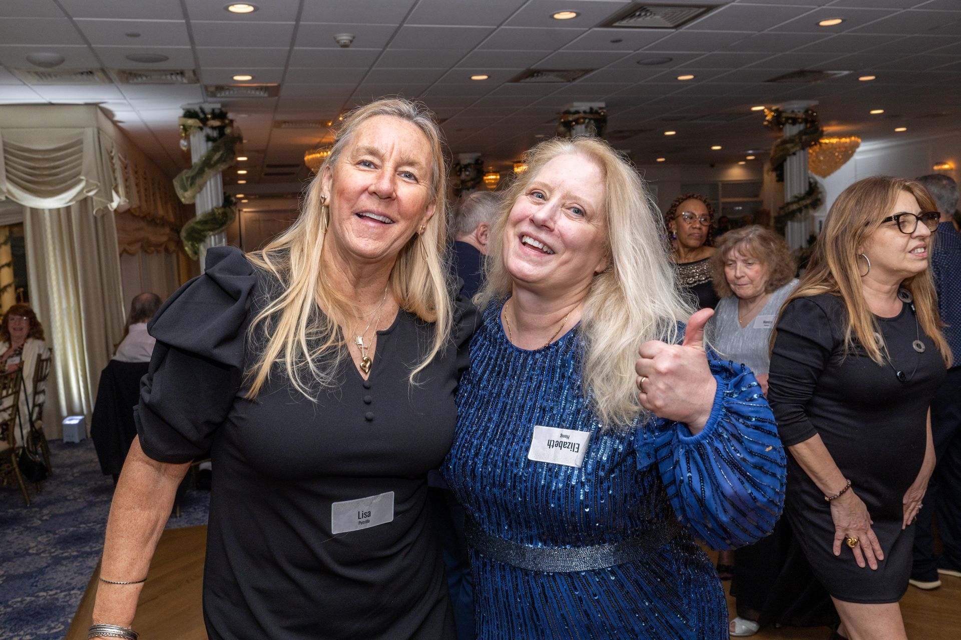 Two women smiling, posing at a party. One gives a thumbs-up. Other guests are visible in the background.