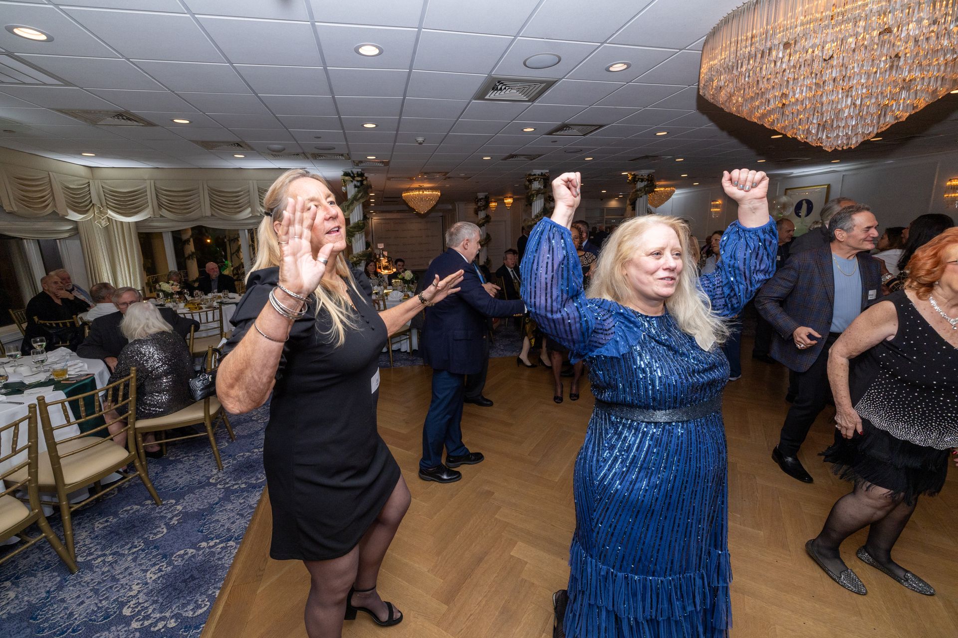 People dancing at a party in a decorated ballroom; one woman in a blue sequined dress.