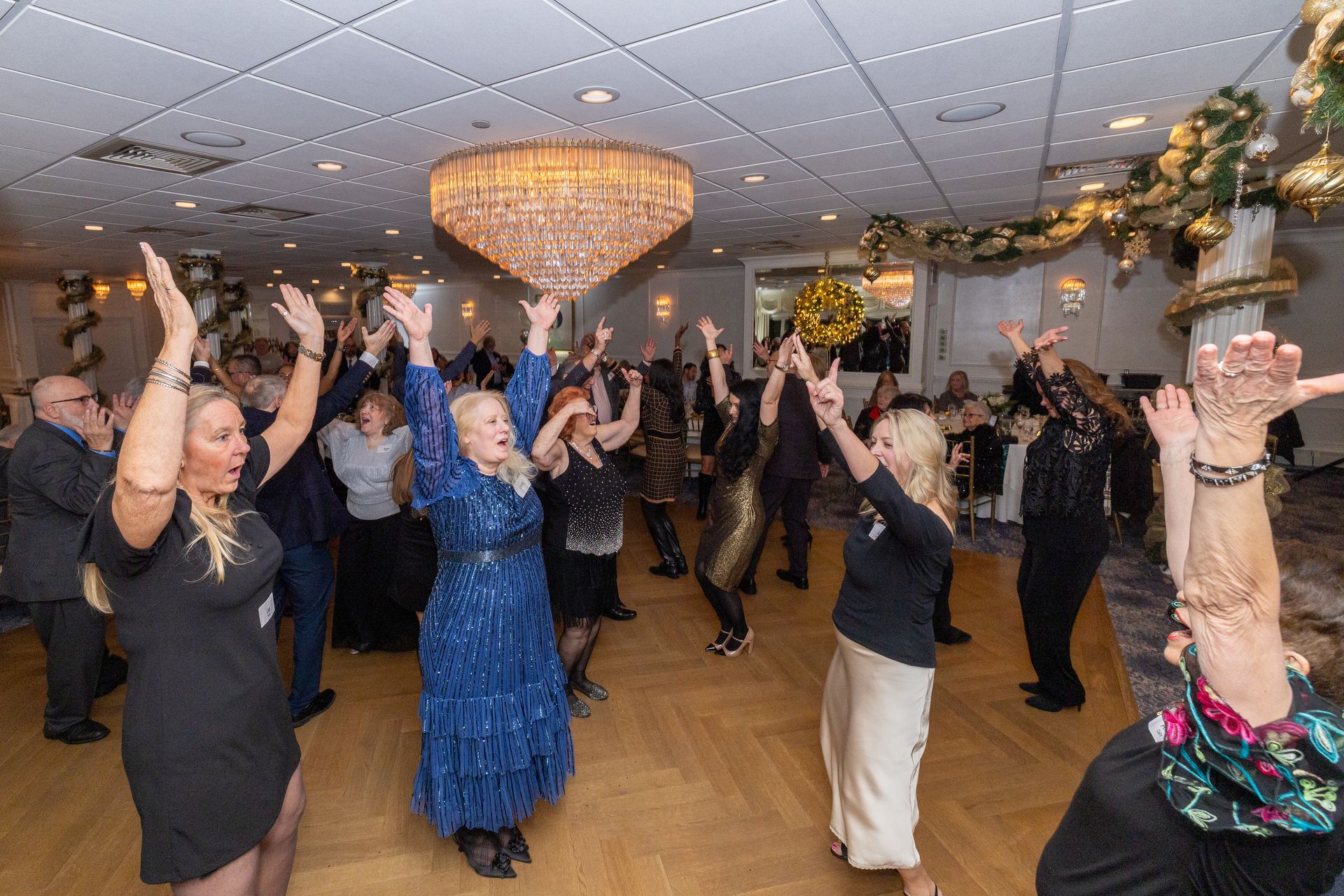 People dancing with raised arms at a festive event. Ballroom with chandelier and gold decor.
