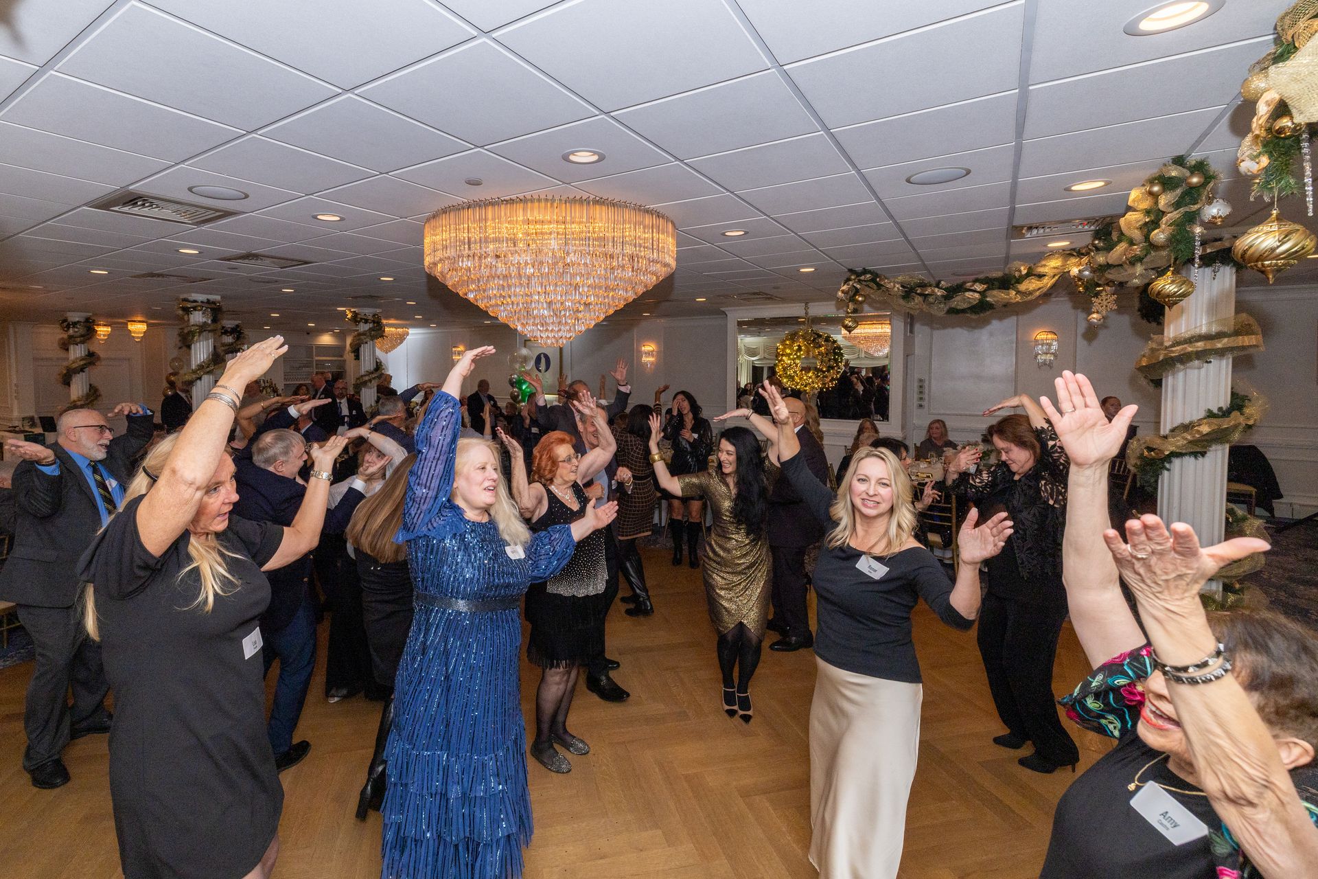 People dancing in a decorated ballroom under a large chandelier. Some have their arms raised.