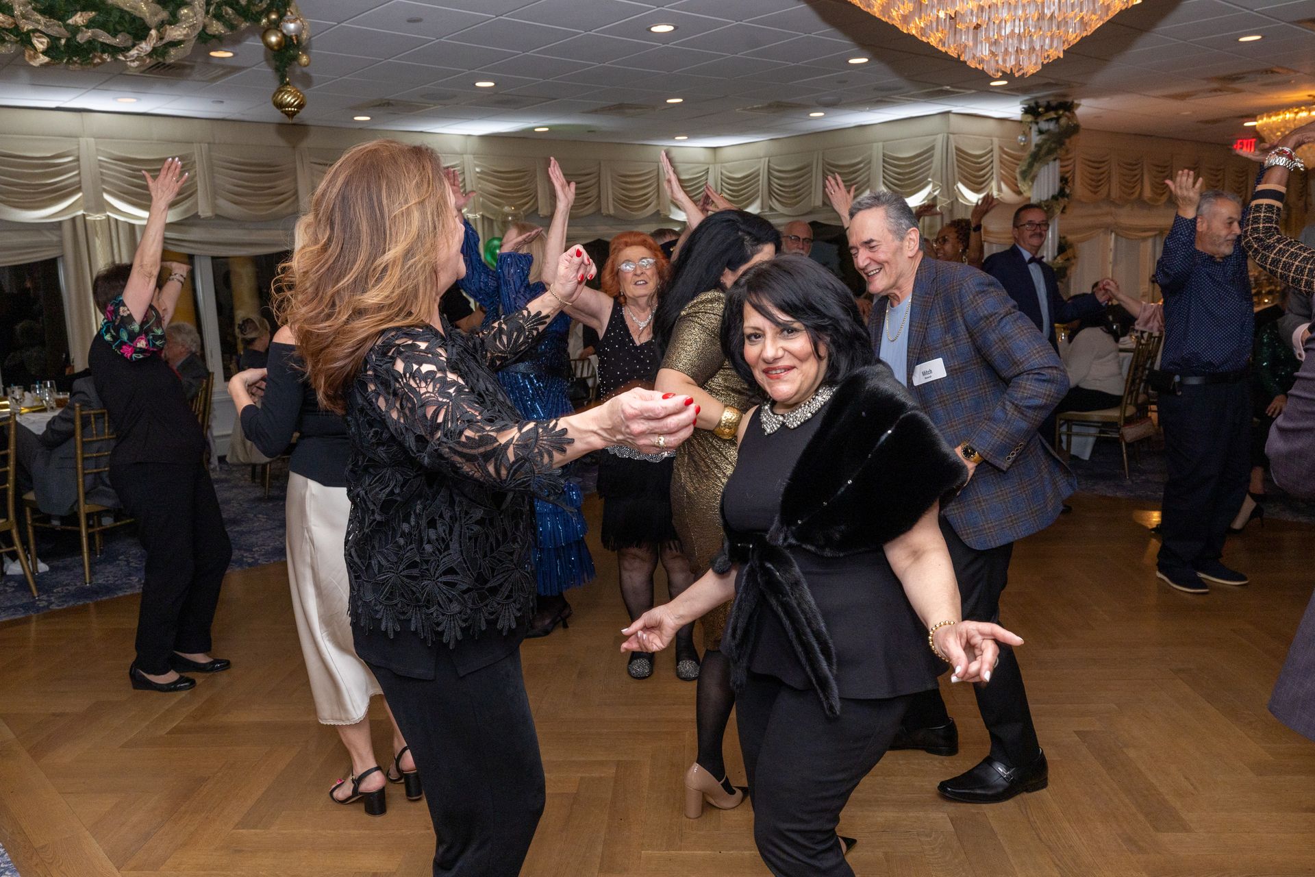 People dancing in a well-lit ballroom; some have arms raised. Festive decor and a chandelier are visible.