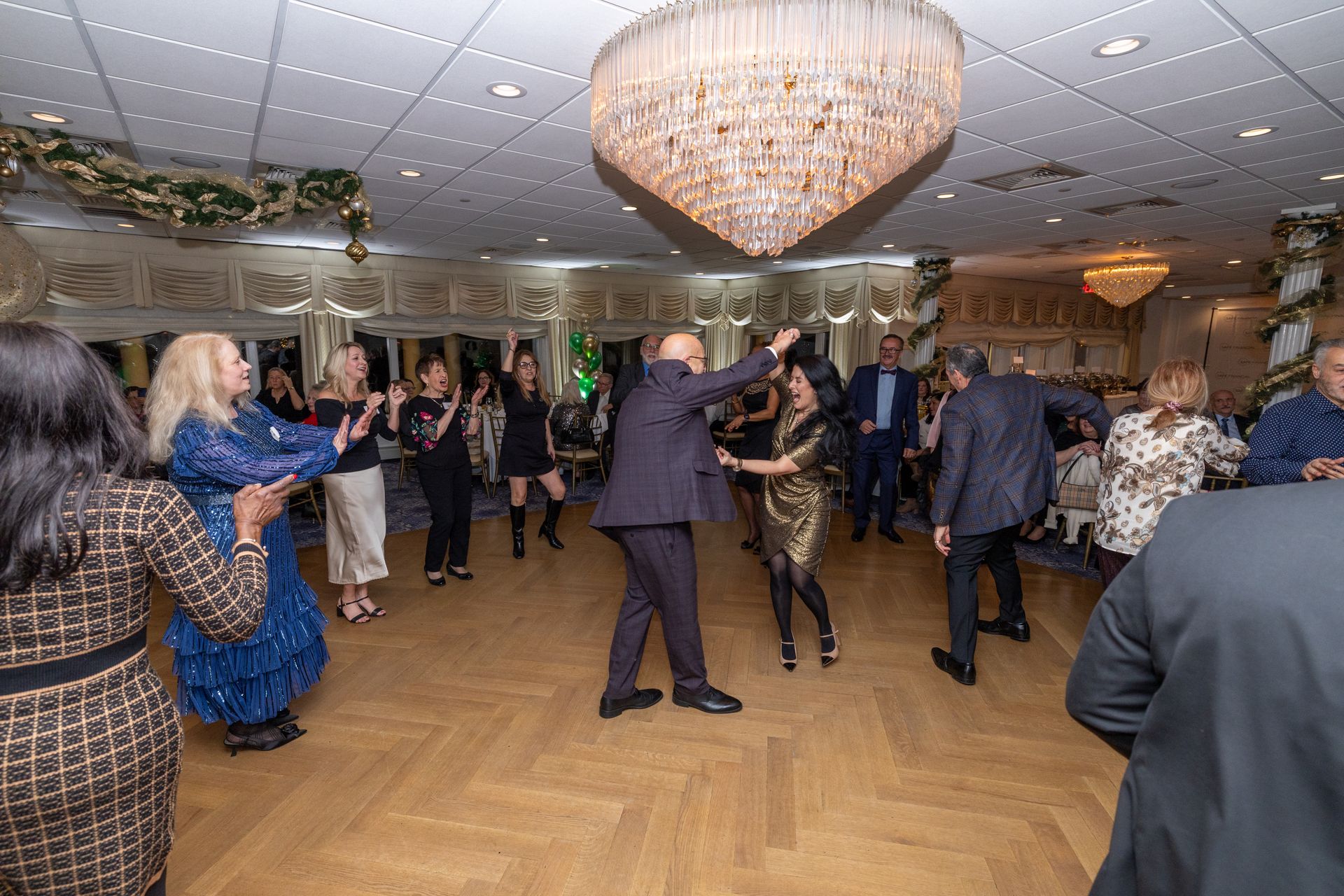 People dancing in a well-lit ballroom with a large chandelier and decorative garlands.