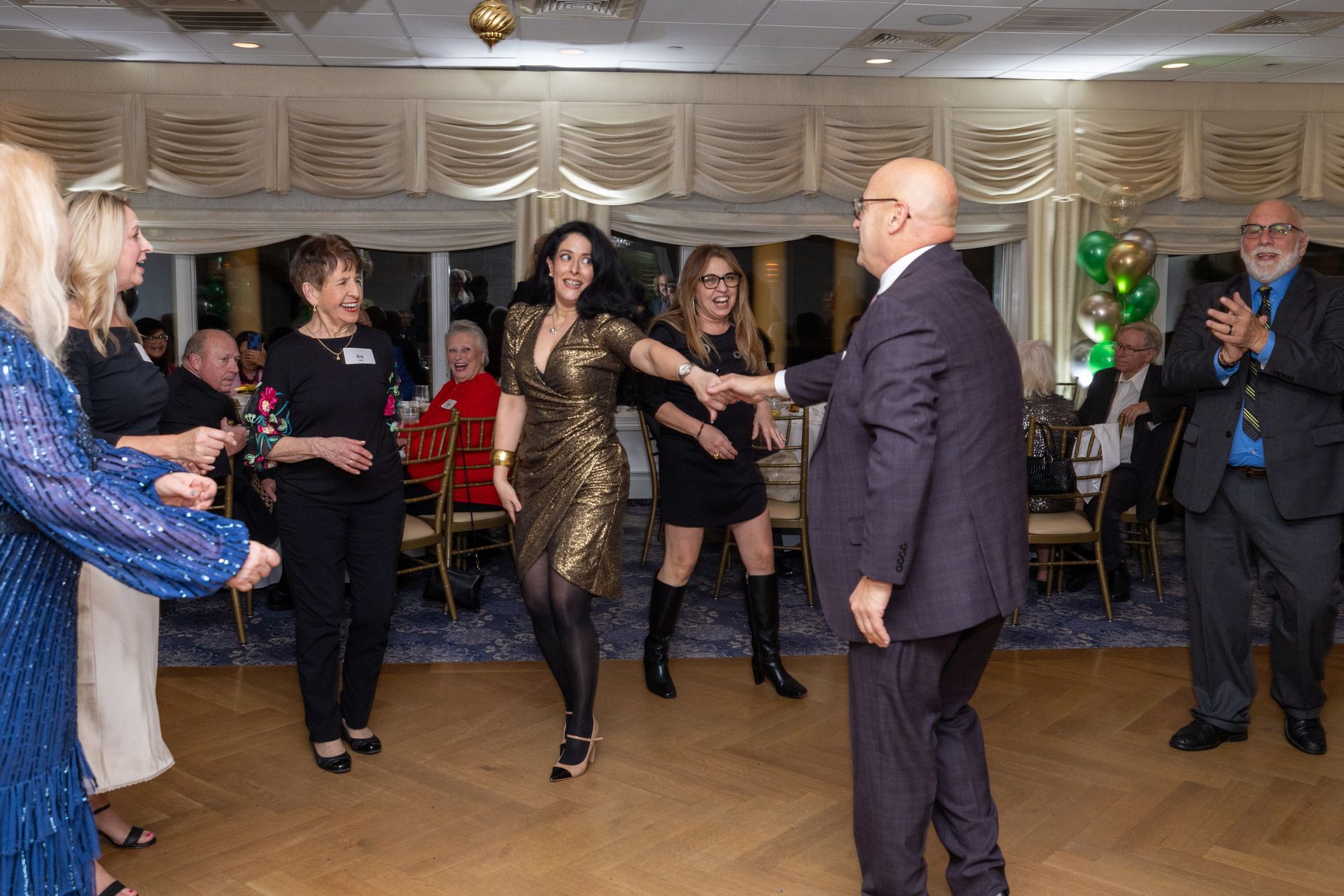 People dancing at a party; woman in gold dress dances with a man in a suit, others clap.
