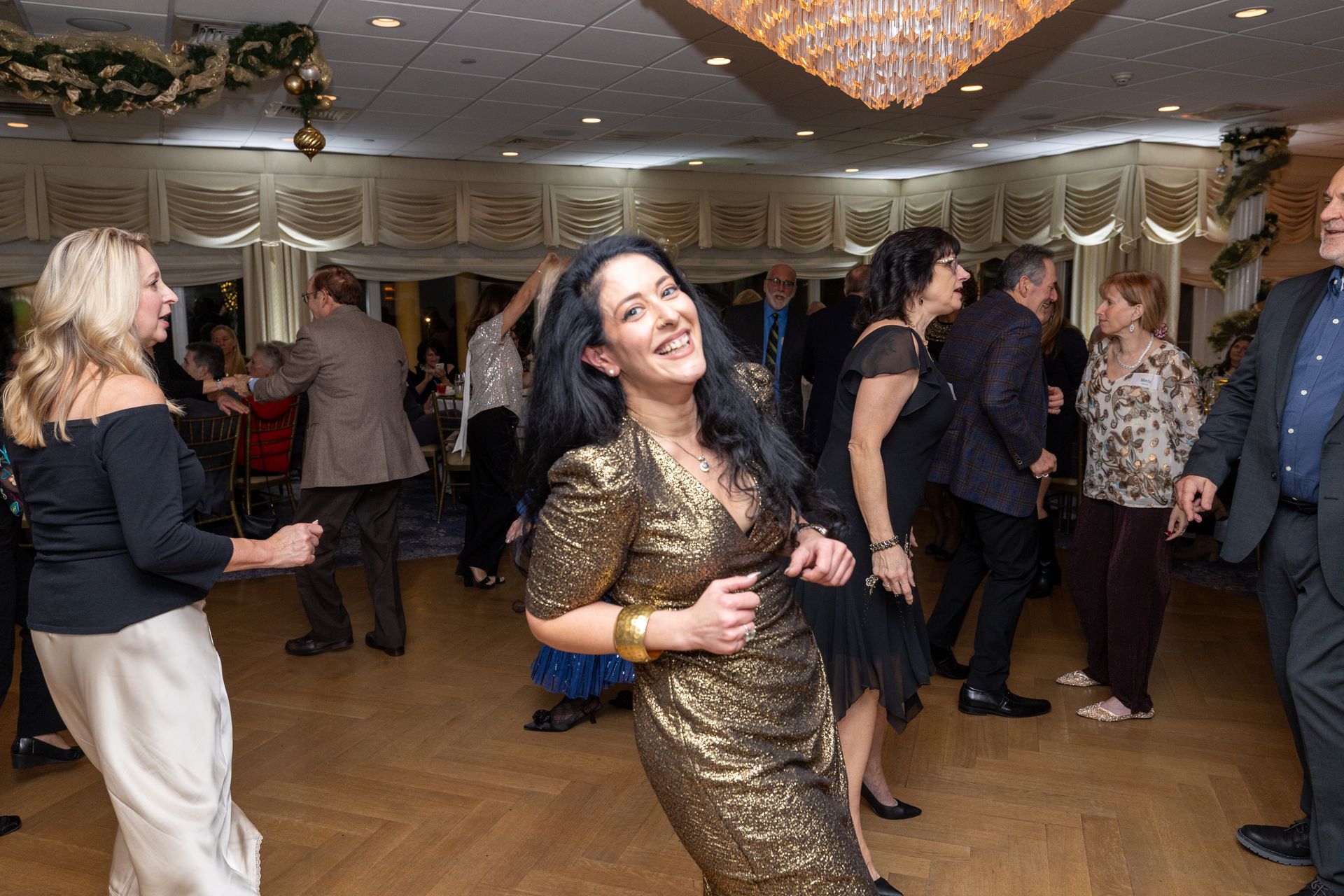 People dancing at a party in a ballroom. Woman in gold dress smiles and dances. Others dance nearby.