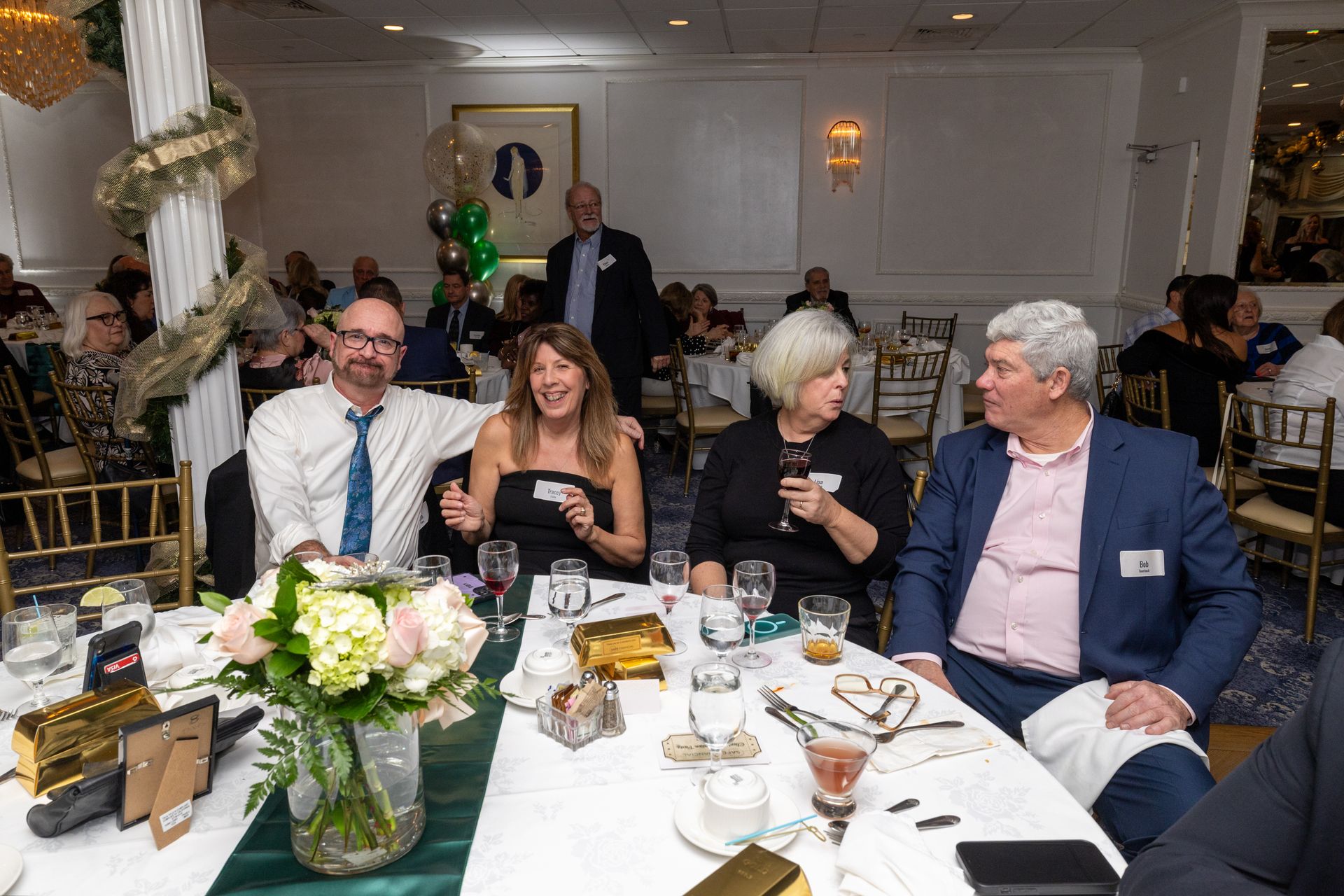 People at a formal event seated at a table, some smiling and conversing. Interior setting with decorations.