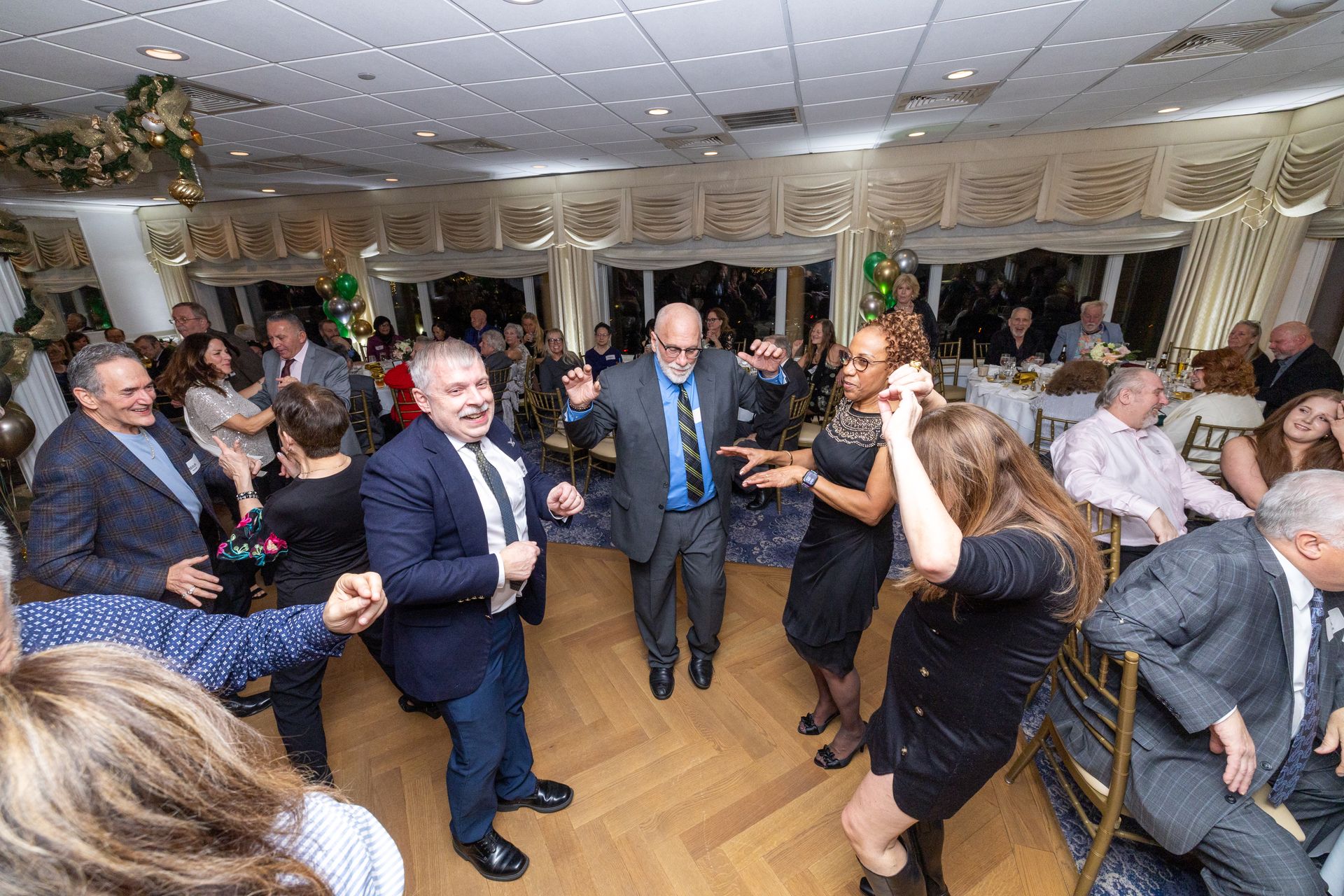 People dancing joyfully at an indoor event with decorations, bright lights, and many guests.