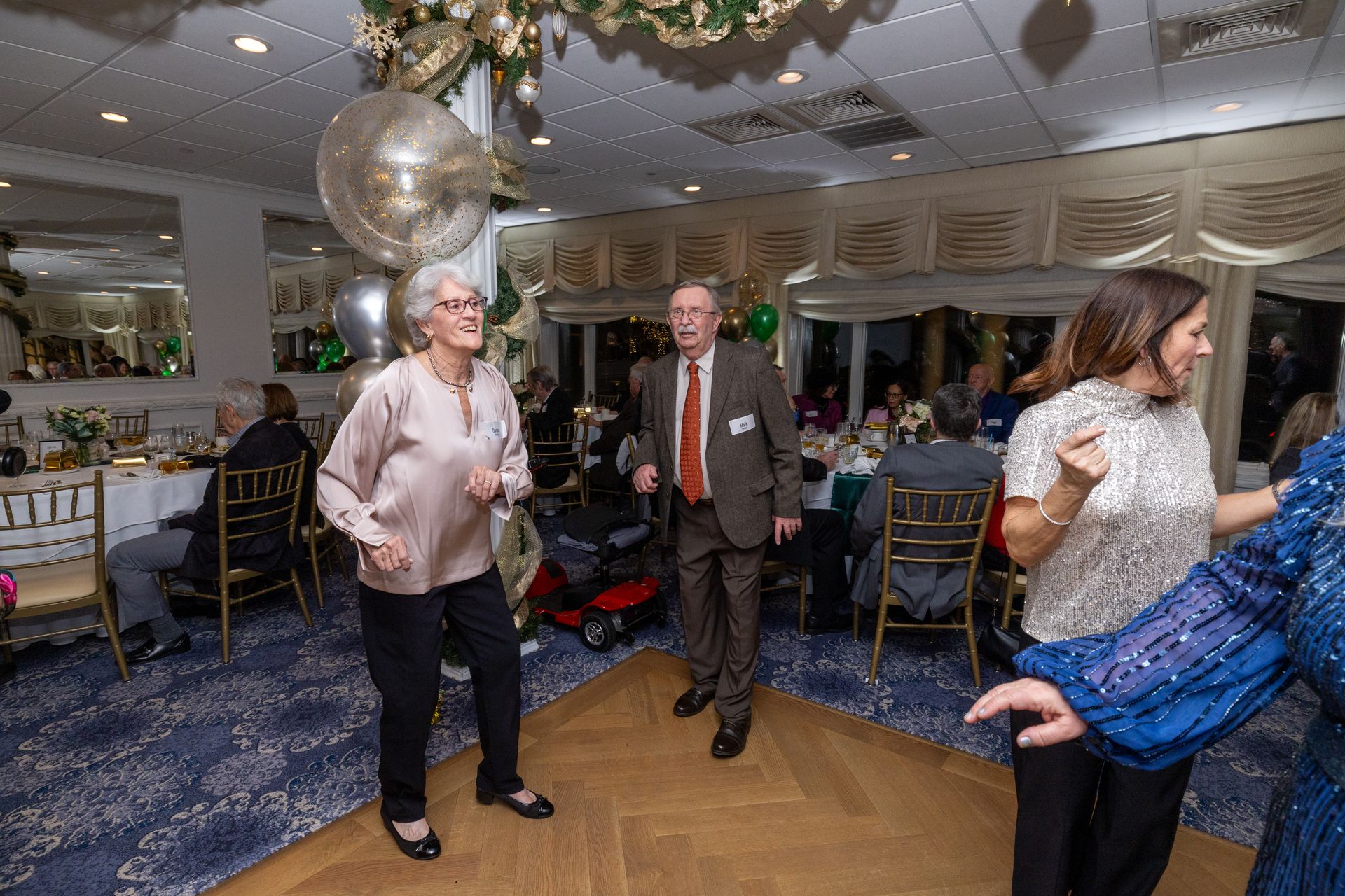 People dancing at a party in a decorated room with tables and a large window.