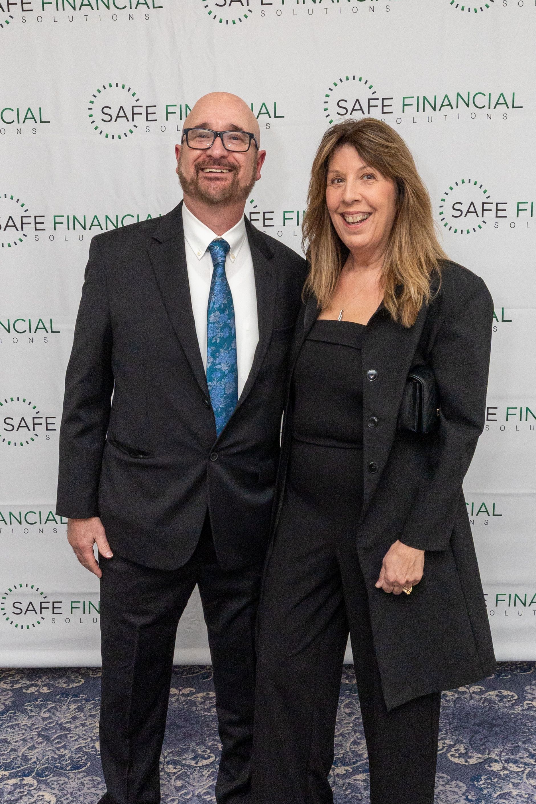 Man and woman posing at an event. The man wears a suit and tie; the woman a pantsuit. Safe Financial logo backdrop.