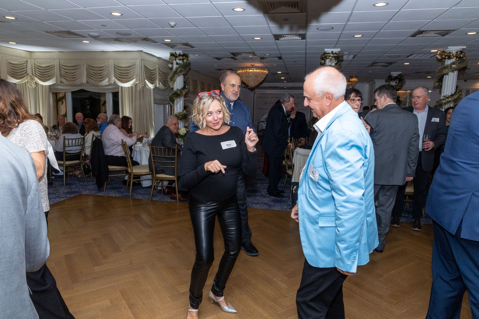 People dancing at a party in a decorated ballroom. A woman in black pants and a man in a blue jacket are central.