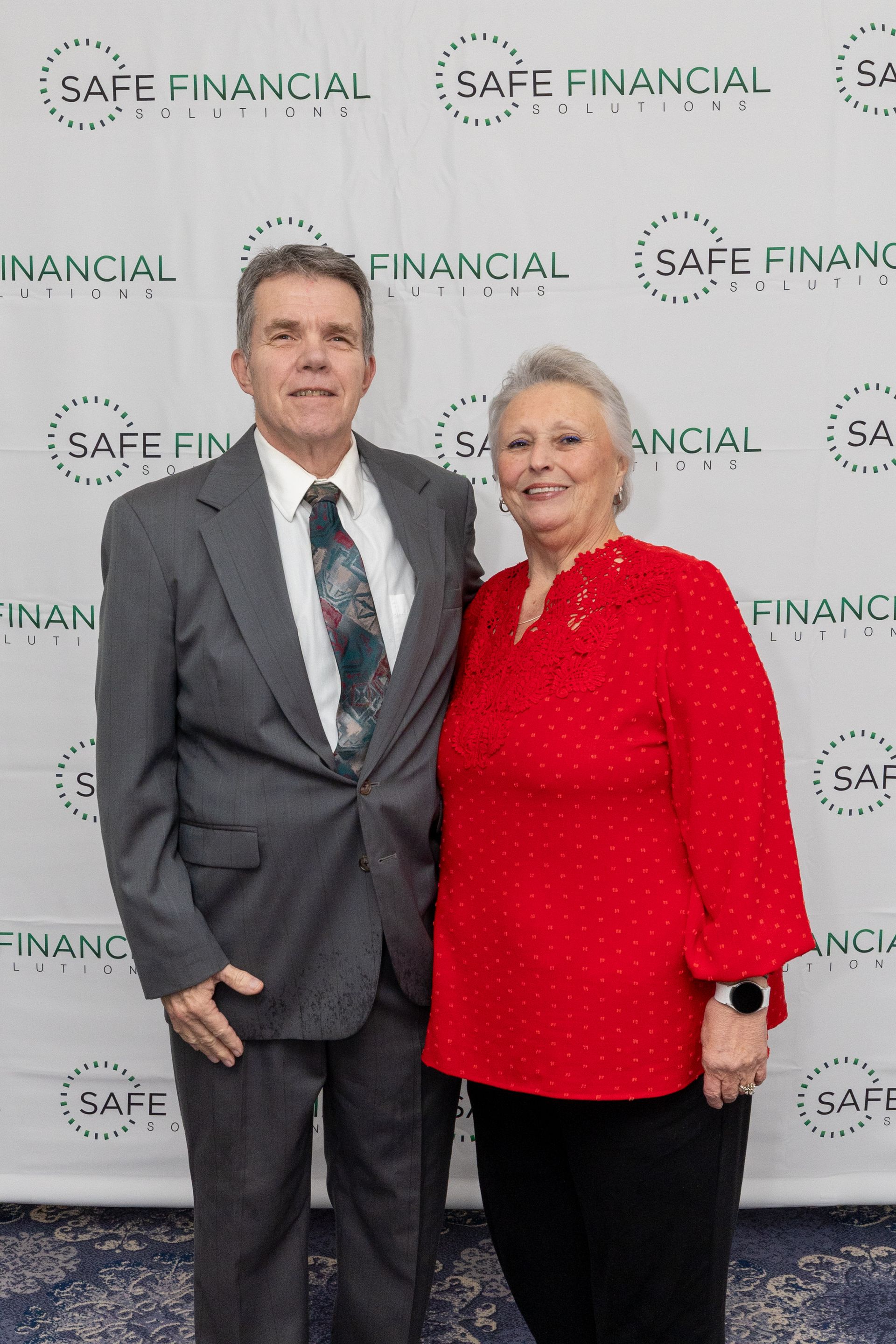 A man and woman posing for a photo. The woman wears a red shirt; the man wears a suit. The backdrop reads 