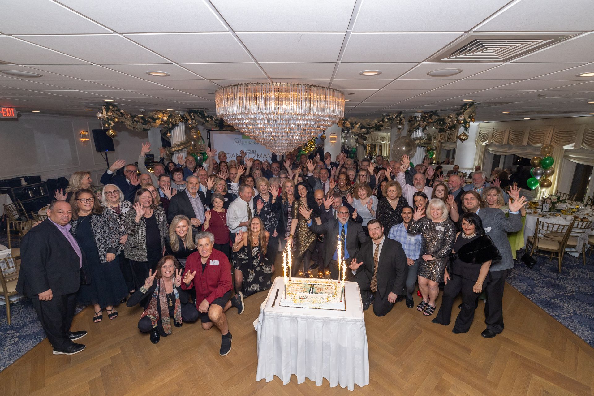 Large group of people gathered around a cake with sparklers; celebratory event in a decorated room.