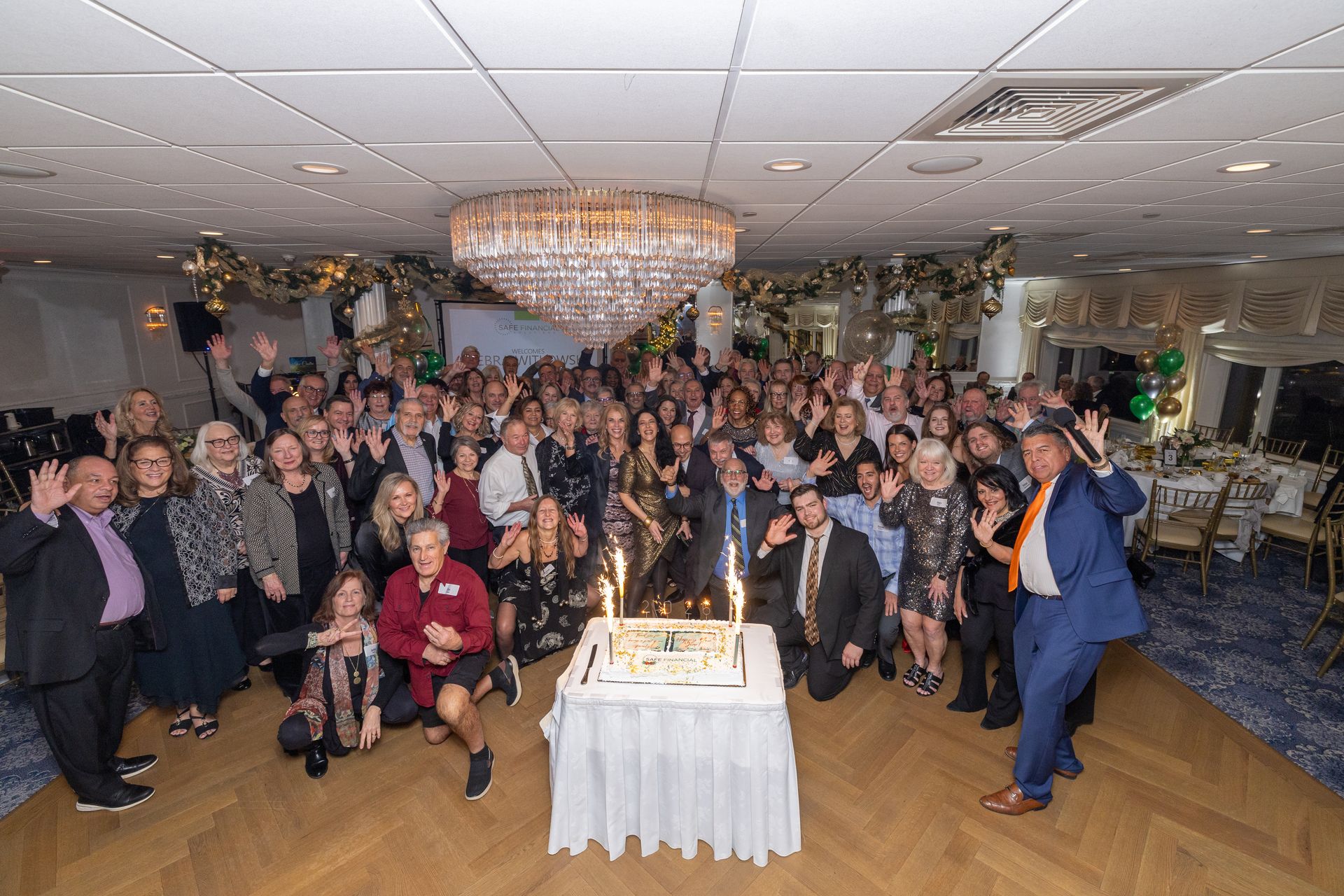 Large group celebrates around a cake with sparklers in a decorated room with a chandelier.
