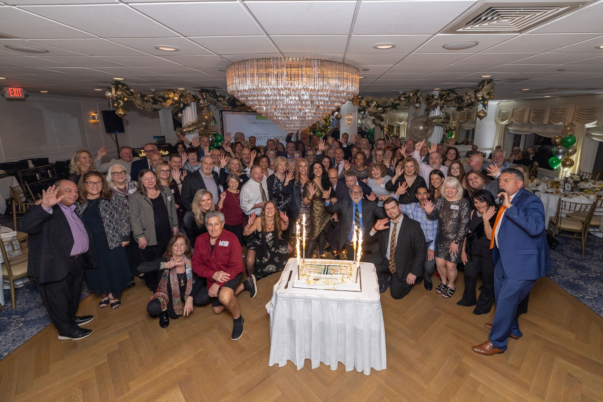 Large group poses around a decorated cake in a banquet hall, celebrating.