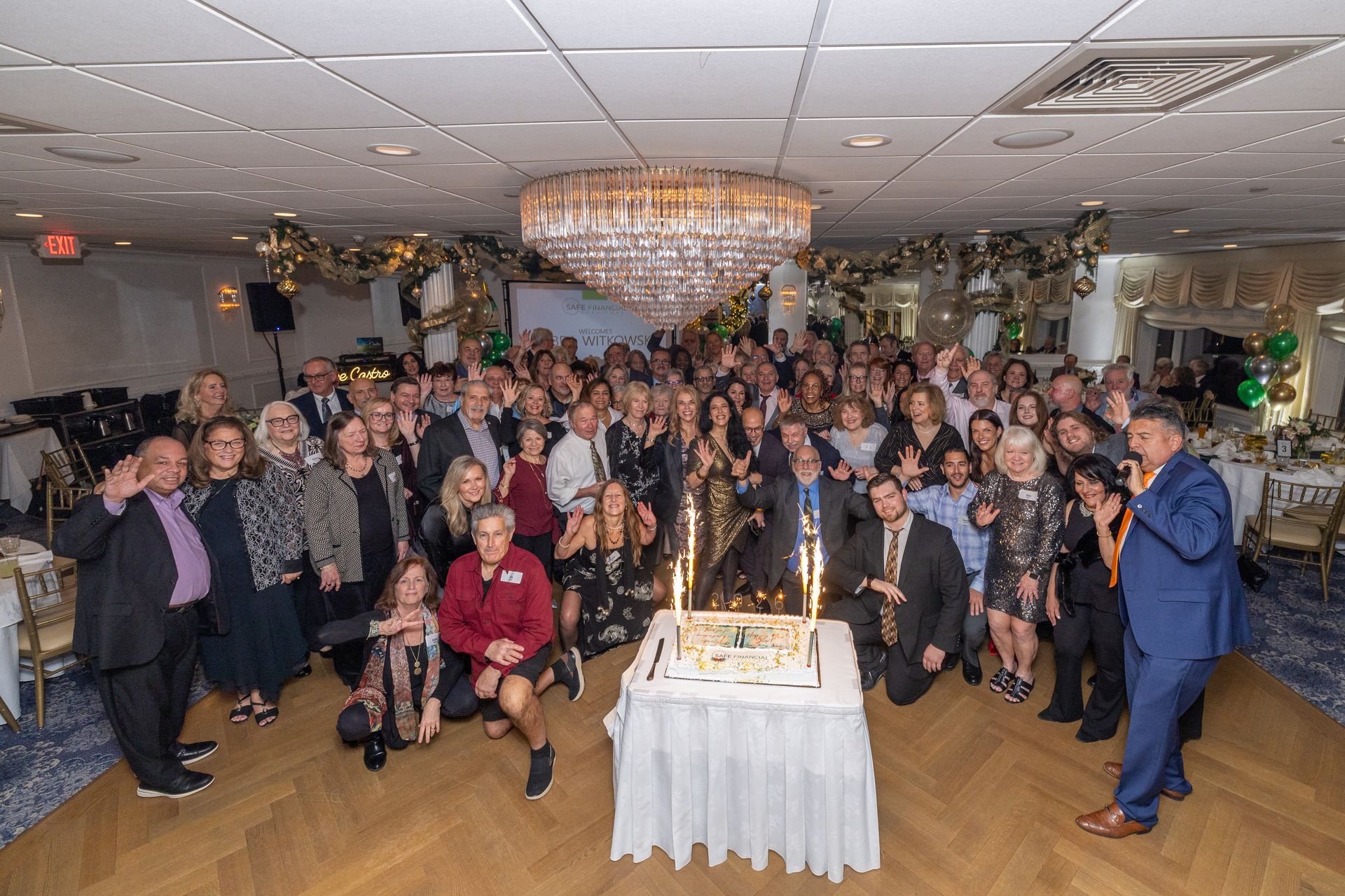Large group of people gathered around a decorated cake at an event with gold and green accents.