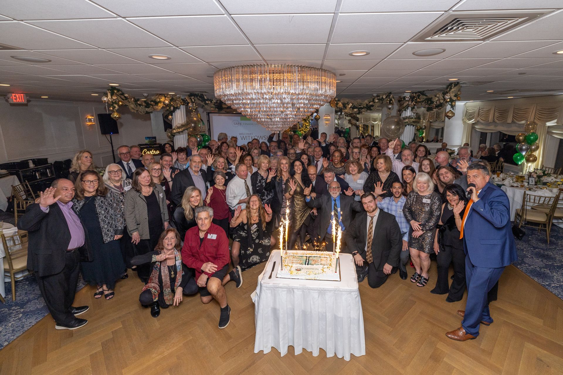 Group of people at a celebration around a cake with sparklers. Room with chandelier, decor, and tables.