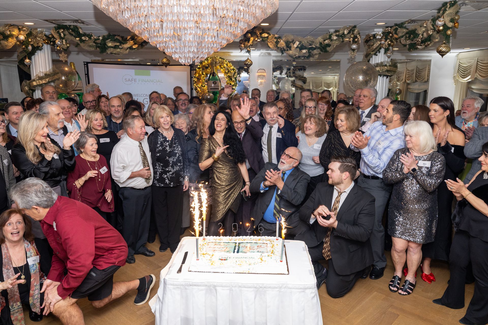 A group celebrates around a cake with sparklers. People clap and smile in an ornate room with a chandelier.