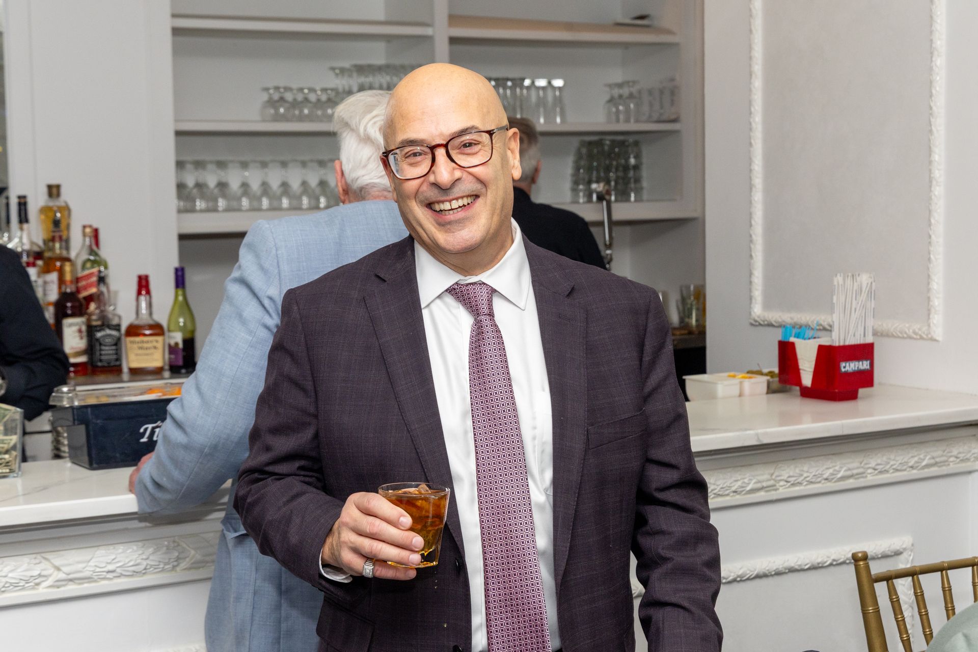 Man in blazer smiles, holding drink at a bar with bottles and glassware.