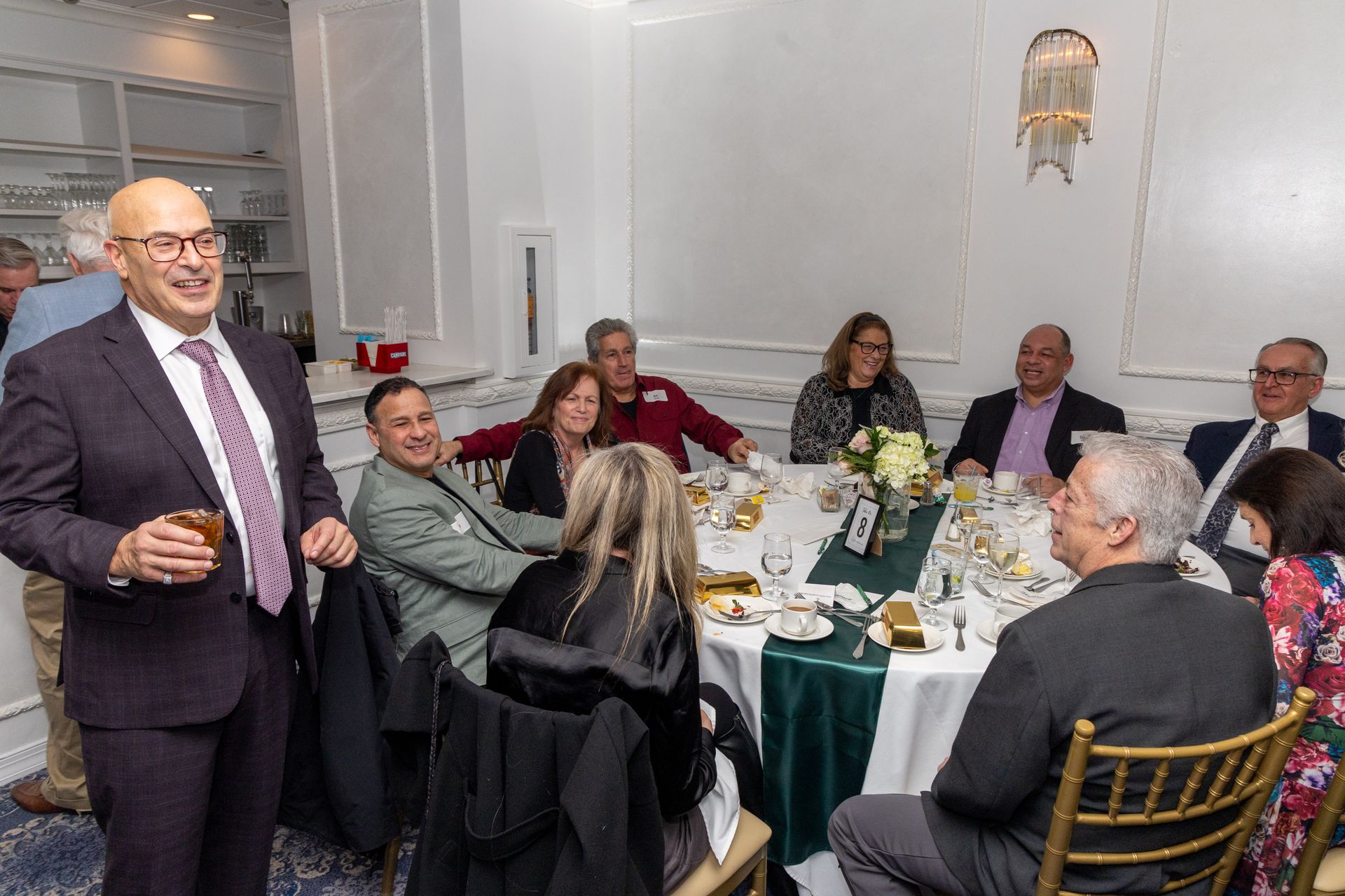 Group of people at a formal event, some seated at a table with a centerpiece, others standing and socializing.